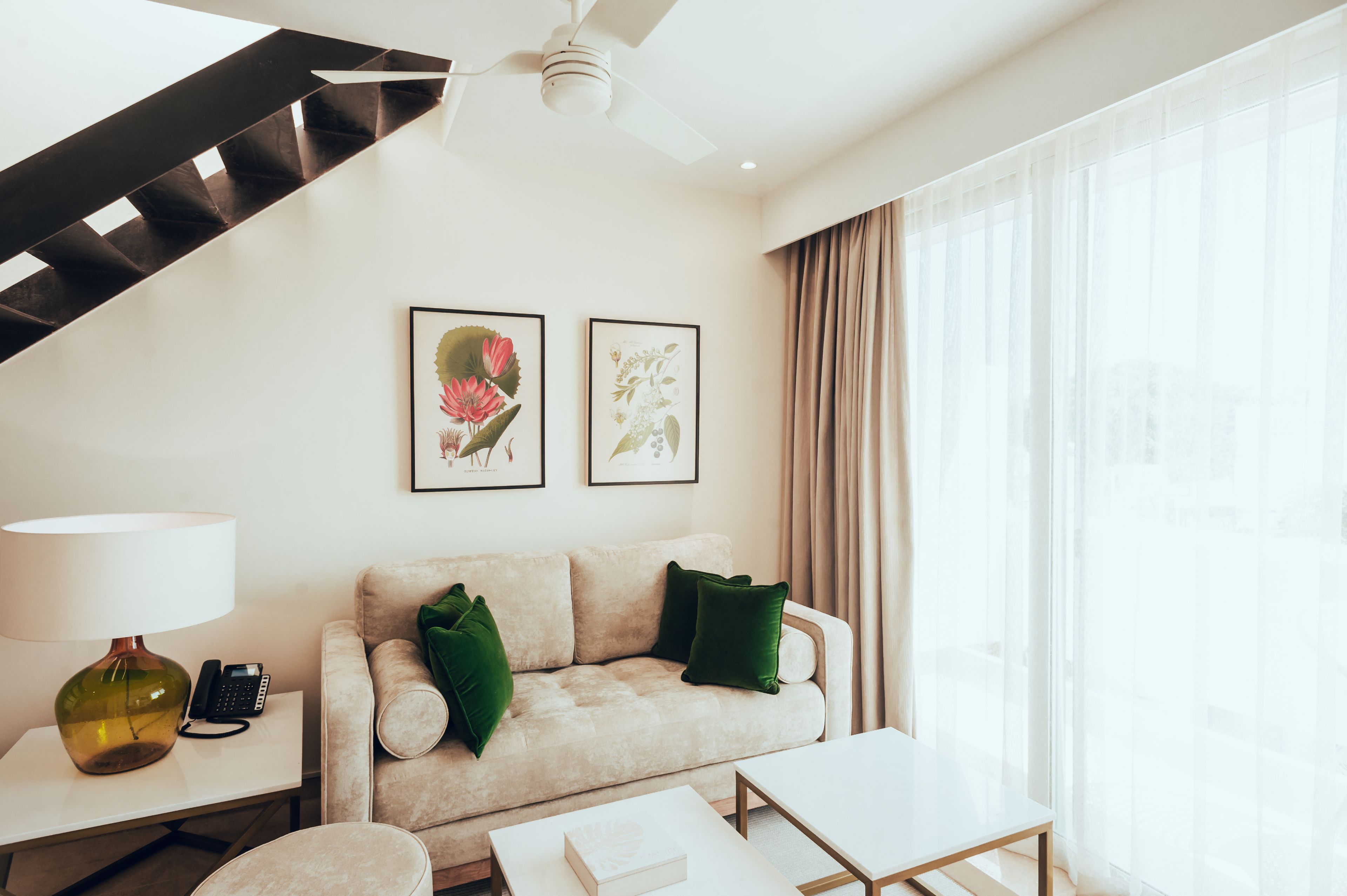 A modern hotel room with a staircase, a sofa with green cushions, a side table with a lamp, and framed botanical artwork on the wall.