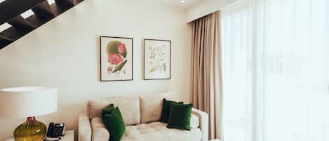 A modern hotel room with a staircase, a sofa with green cushions, a side table with a lamp, and framed botanical artwork on the wall.