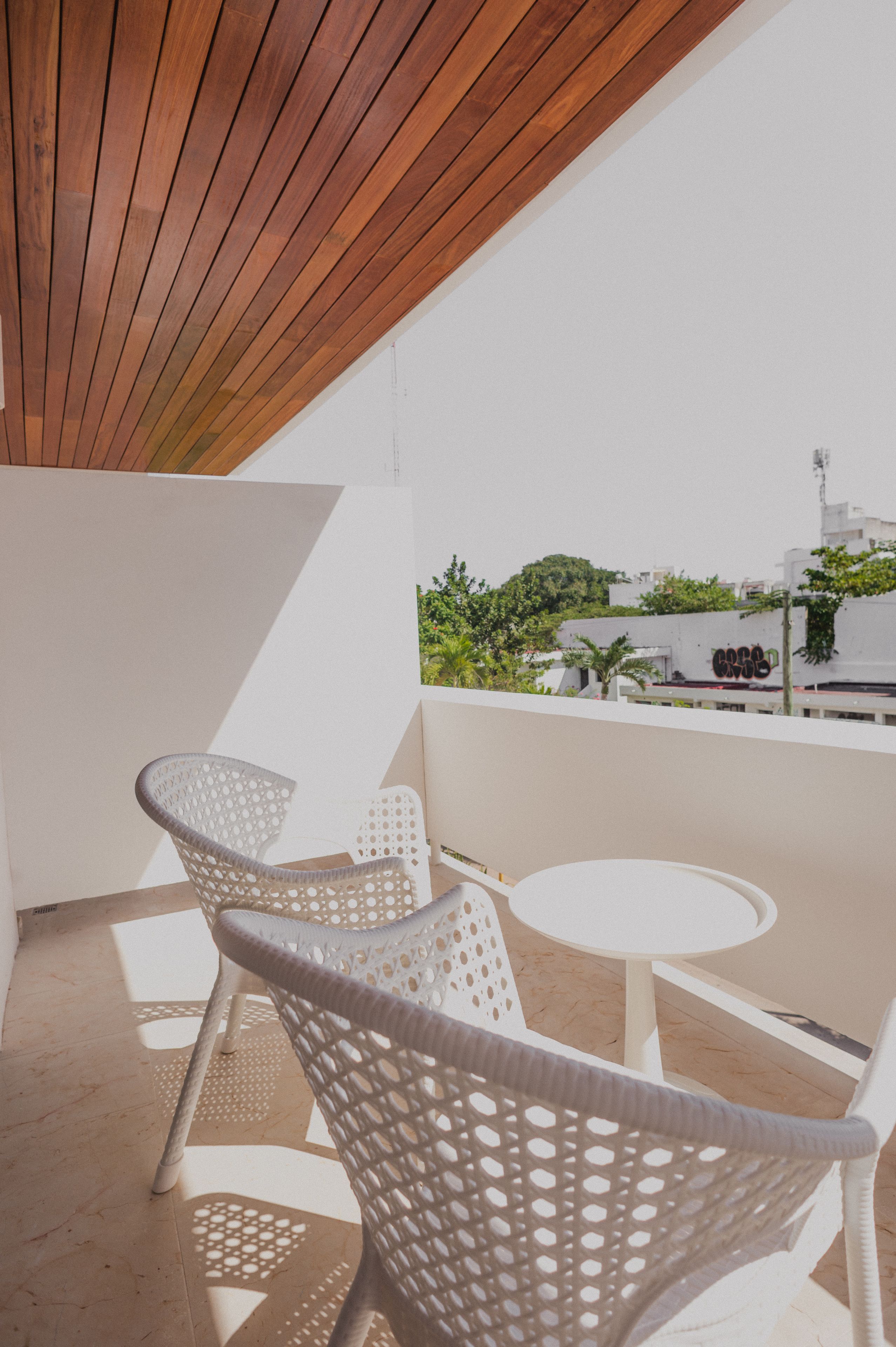 A balcony with white wicker chairs and a small round table, overlooking a cityscape with greenery and buildings.