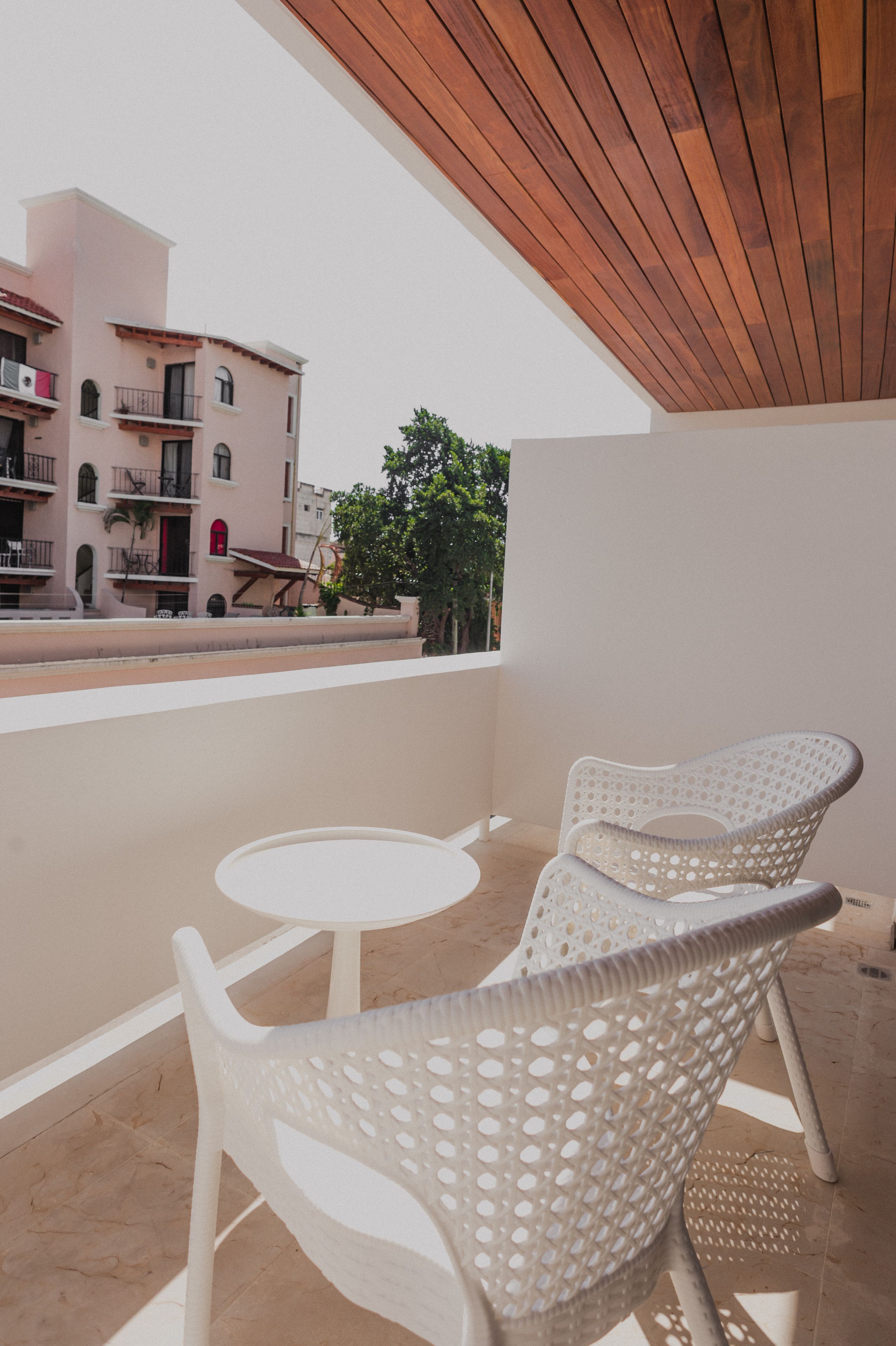A balcony with white plastic chairs and a small round table, overlooking a pink building with balconies and a tree.