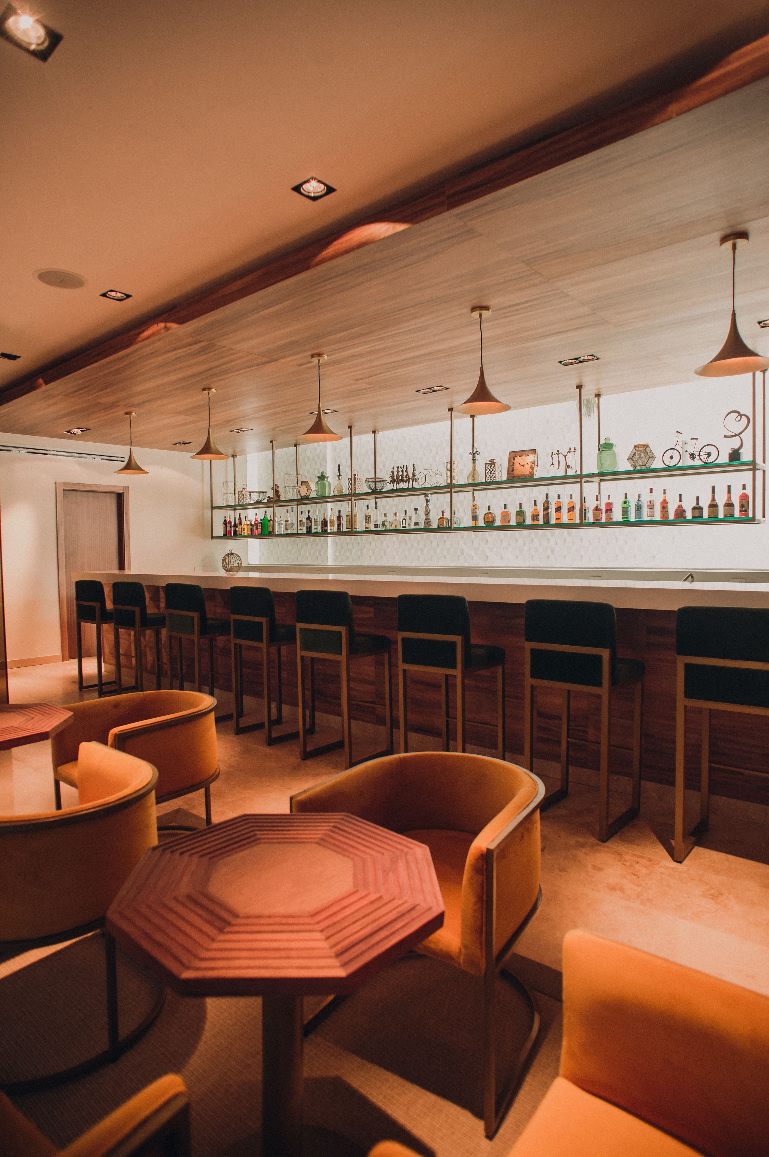 A bar area with wooden bar stools, a long counter with bottles, and pendant lighting.