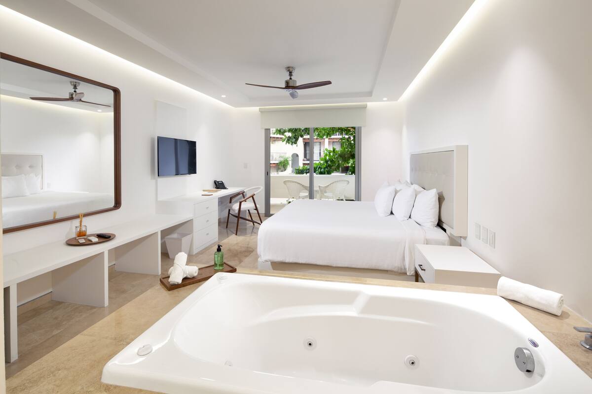 A modern hotel room with a large bed, a separate desk, a vanity with a mirror, a large bathtub, and a balcony with a view of greenery.