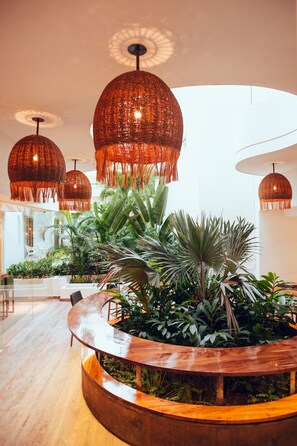Un interior moderno con una mesa redonda de madera rodeada de abundante follaje, grandes lámparas colgantes de mimbre y un ambiente luminoso y espacioso.