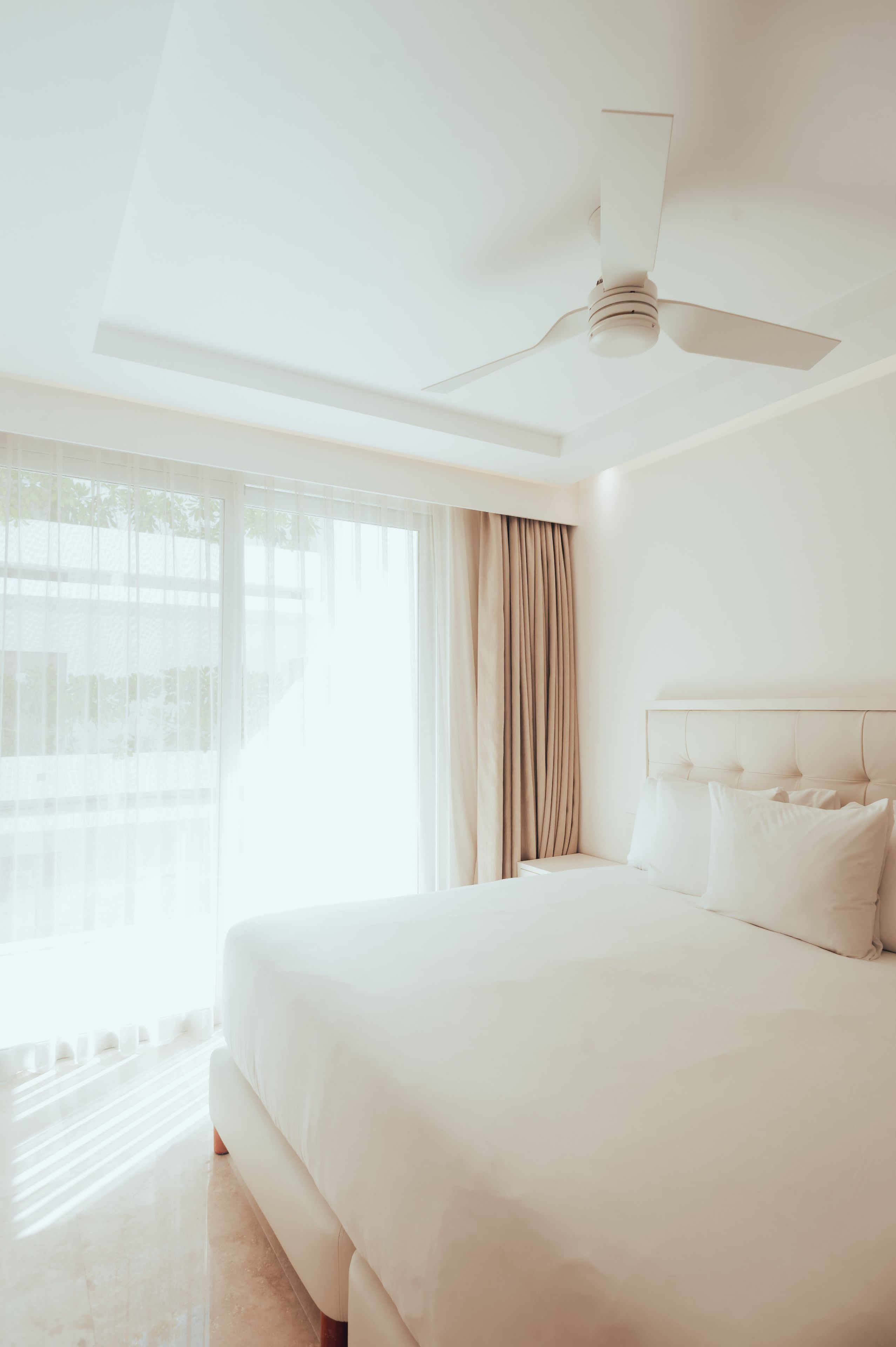 A hotel room with a large bed, a ceiling fan, and a view of the outdoors through large windows.