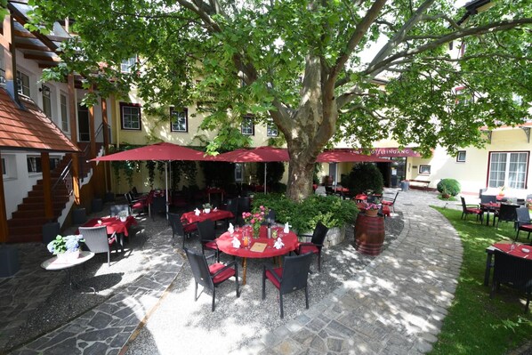 Un espace extérieur aménagé pour les repas, avec des tables et des chaises, protégé par des parasols rouges et entouré d’arbres.