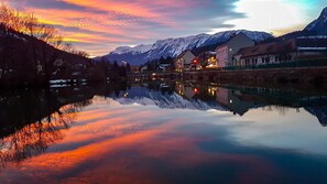 A calm river reflecting a sunset with snow-capped mountains in the background and buildings along the riverbank.