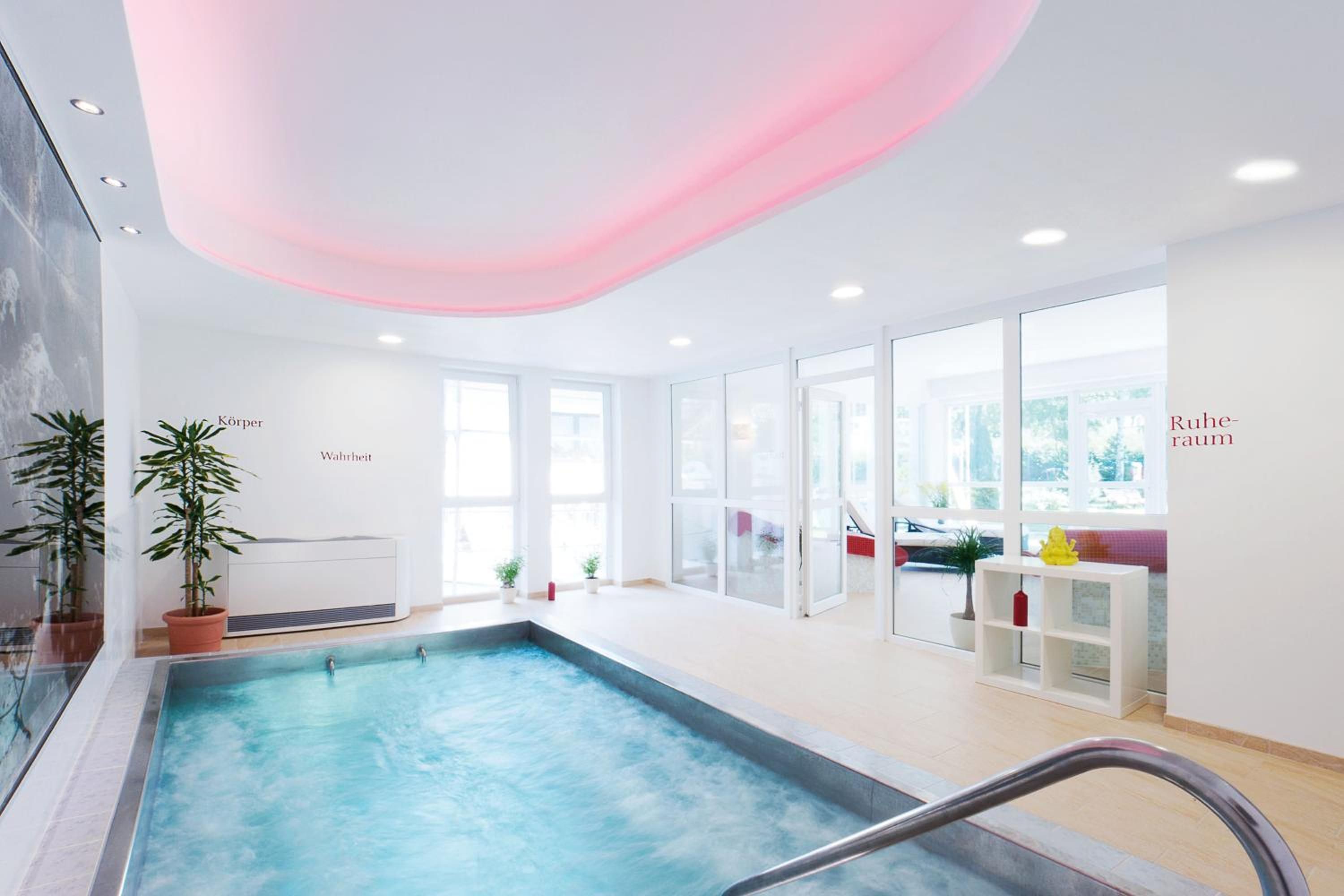 An indoor swimming pool with clear glass walls, potted plants, and a ceiling with a pink light.