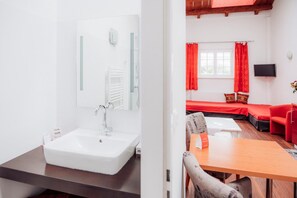 Une salle de bain moderne avec un lavabo et un miroir blancs, une table en bois et un canapé rouge dans l’espace de vie adjacent.