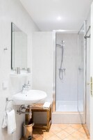 Un baño con un lavamanos blanco, un espejo, una ducha con puerta de vidrio y un taburete de madera.