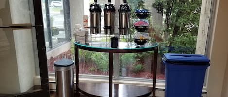 A corner of a room with a glass table holding bottles, a trash bin, and a window with a view of trees and a building.