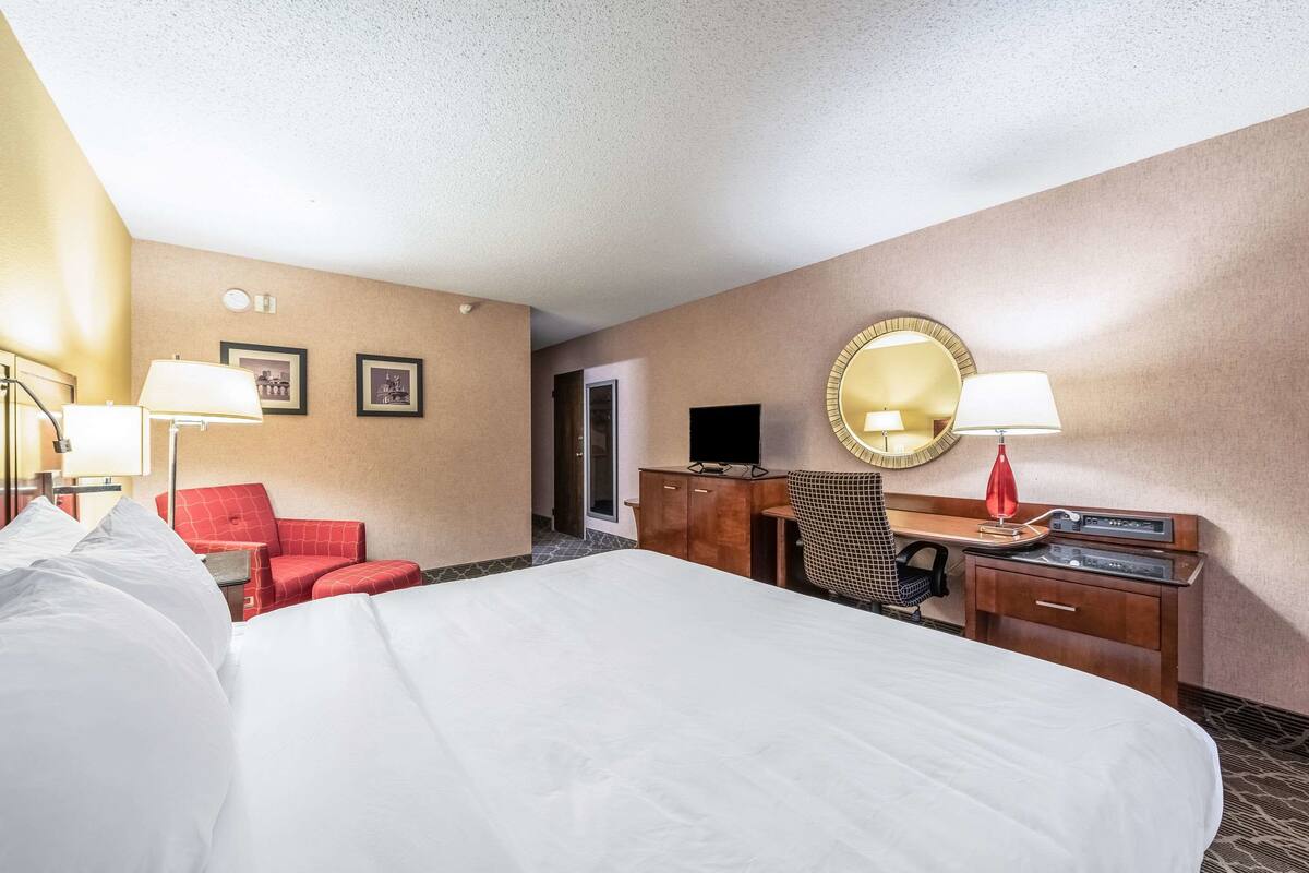 A hotel room with a large bed, a desk with a lamp, a red chair, a television, and a mirror.