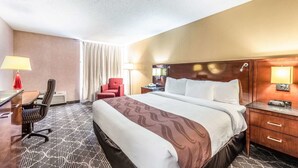 Una habitación de hotel con una cama grande, un escritorio con una silla, un sillón rojo, una mesita de noche con una lámpara y un cabecero de madera.