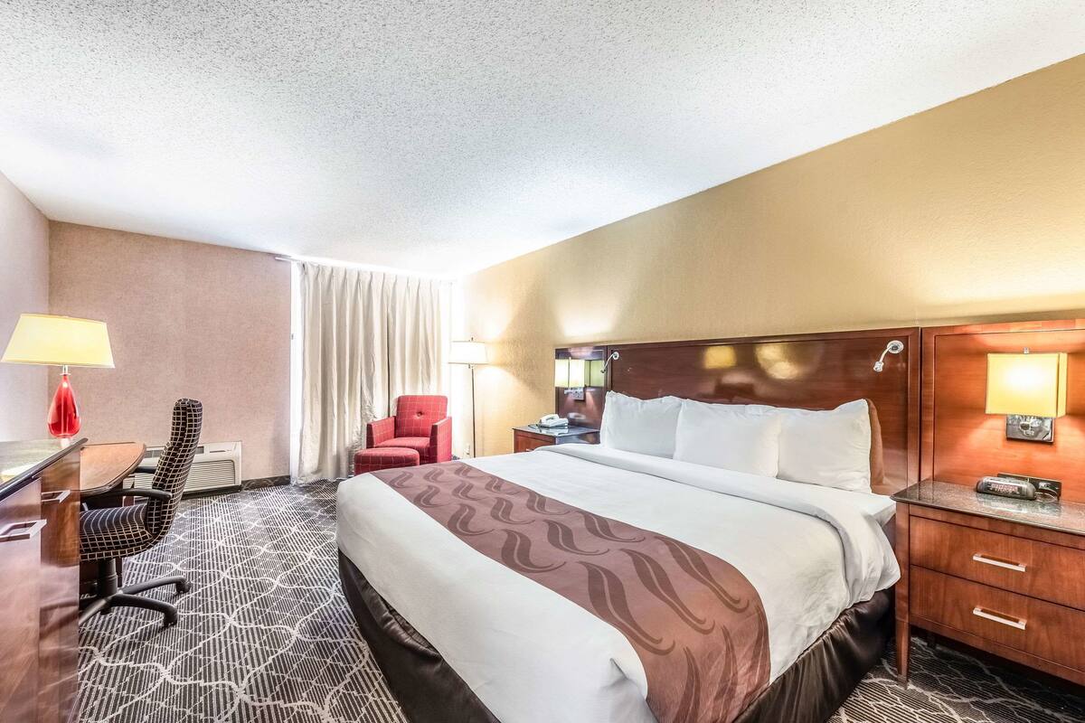 A hotel room with a large bed, a desk with a chair, a red armchair, a nightstand with a lamp, and a wooden headboard.