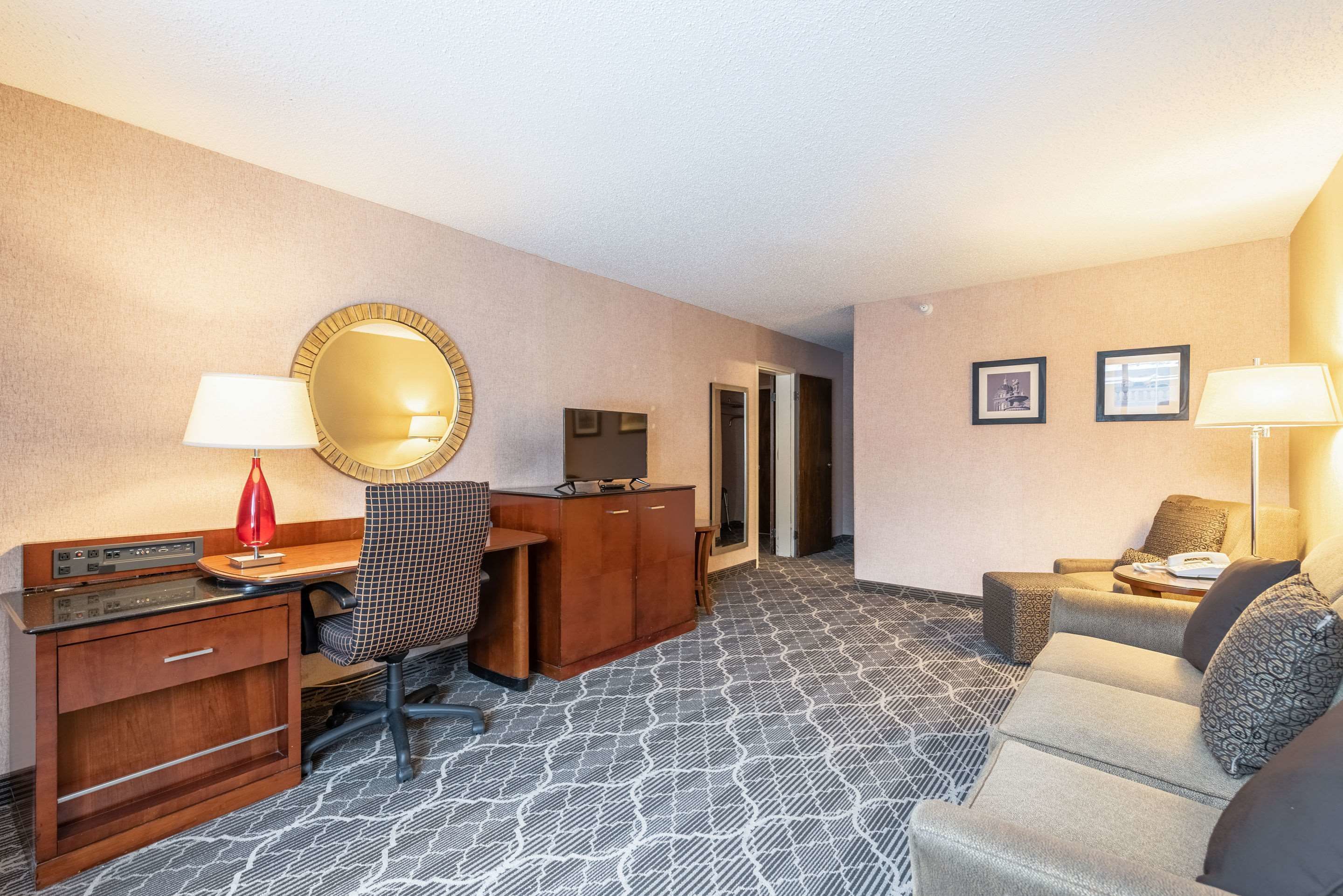 A hotel room with a desk, chair, television, sofa, and patterned carpet.