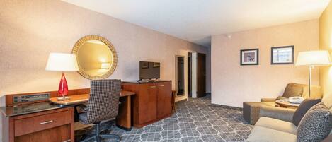 A hotel room with a desk, chair, television, sofa, and patterned carpet.
