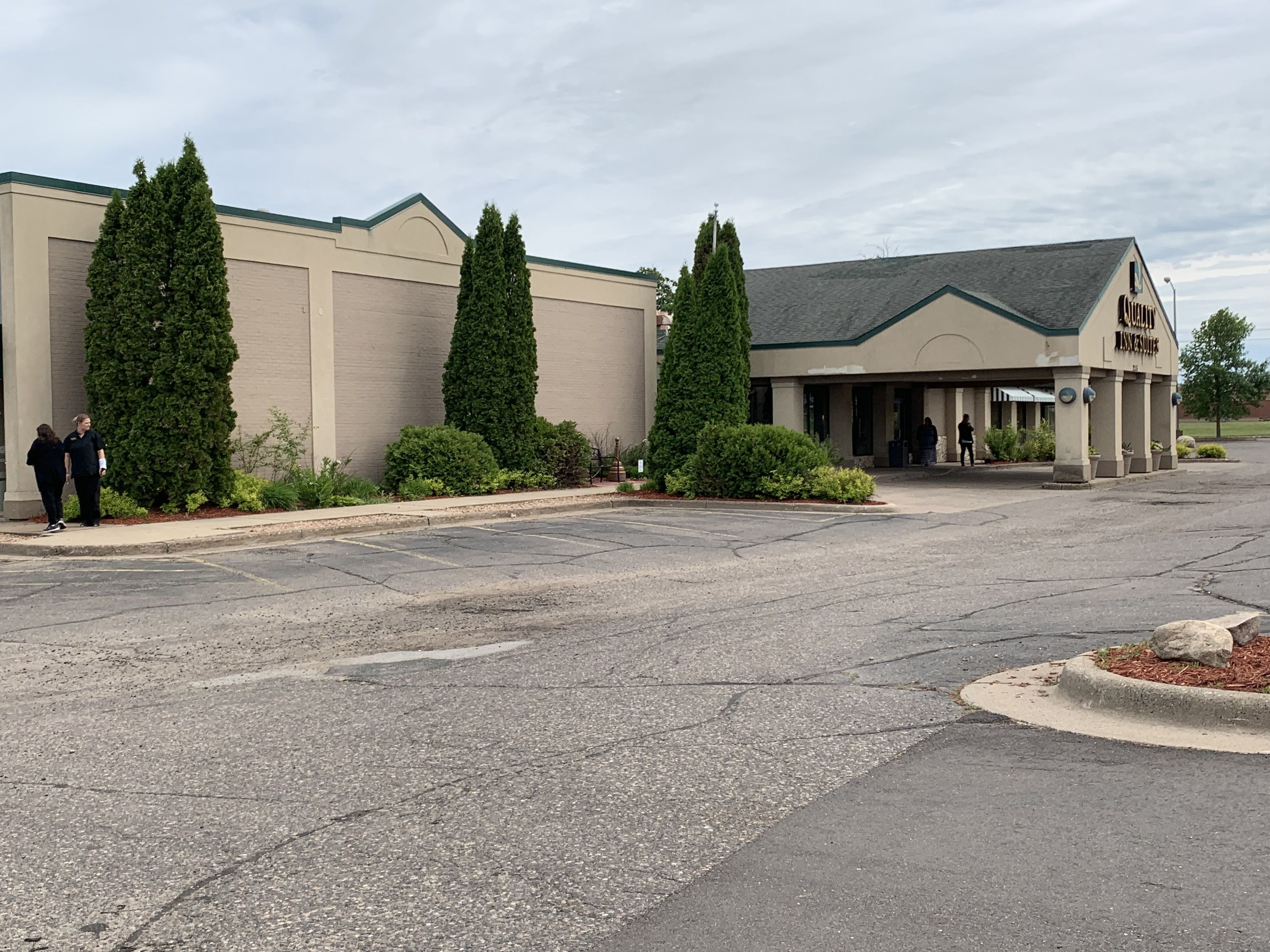 A building with a beige exterior, a green roof, and a sign reading "Quality Inn & Suites." There are three tall, narrow trees in front, and a parking lot with visible cracks.