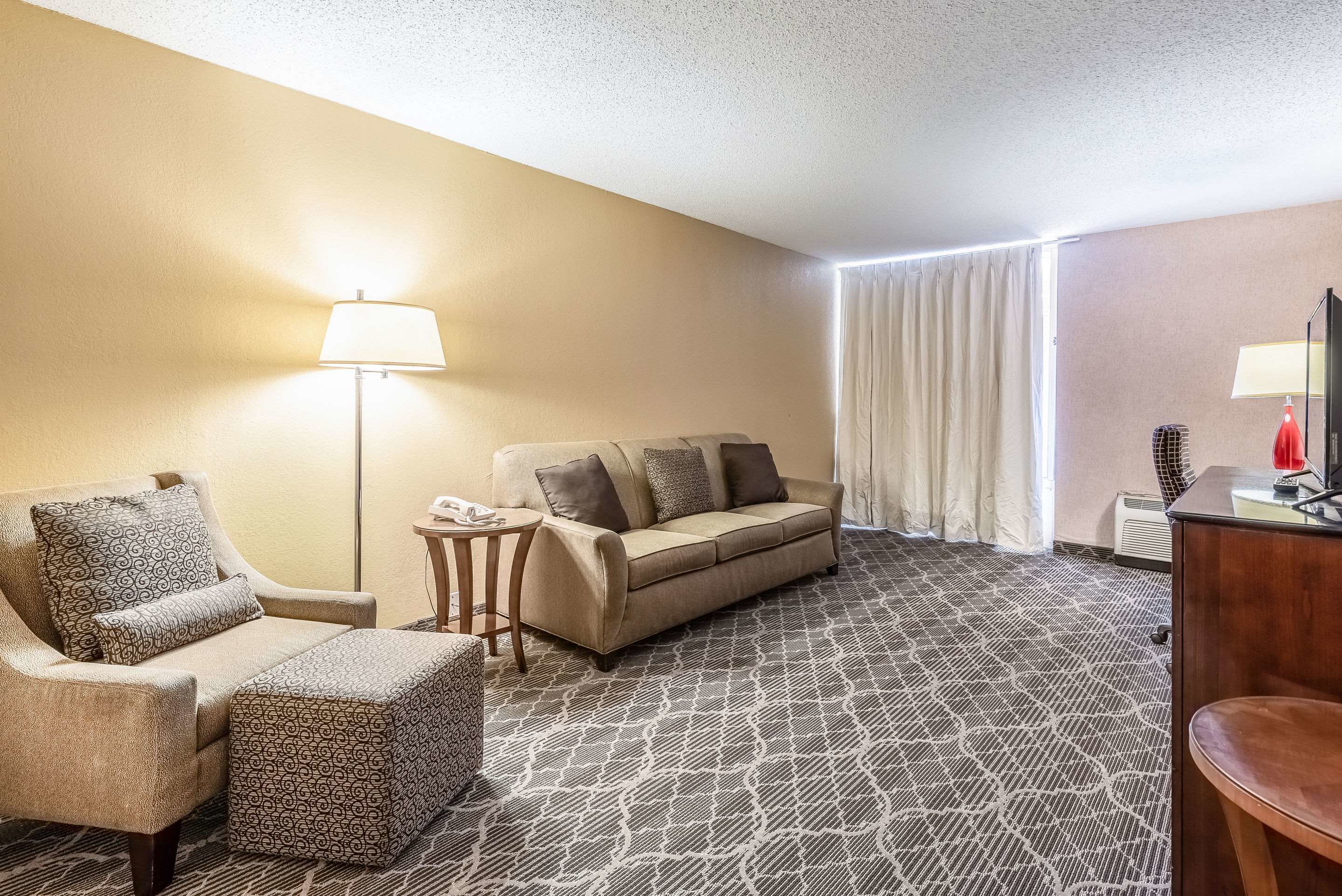 Una habitación de hotel con un sofá color beige, un sillón a juego, un reposapiés estampado, una mesita de noche con un teléfono, una lámpara y un escritorio con un espejo.