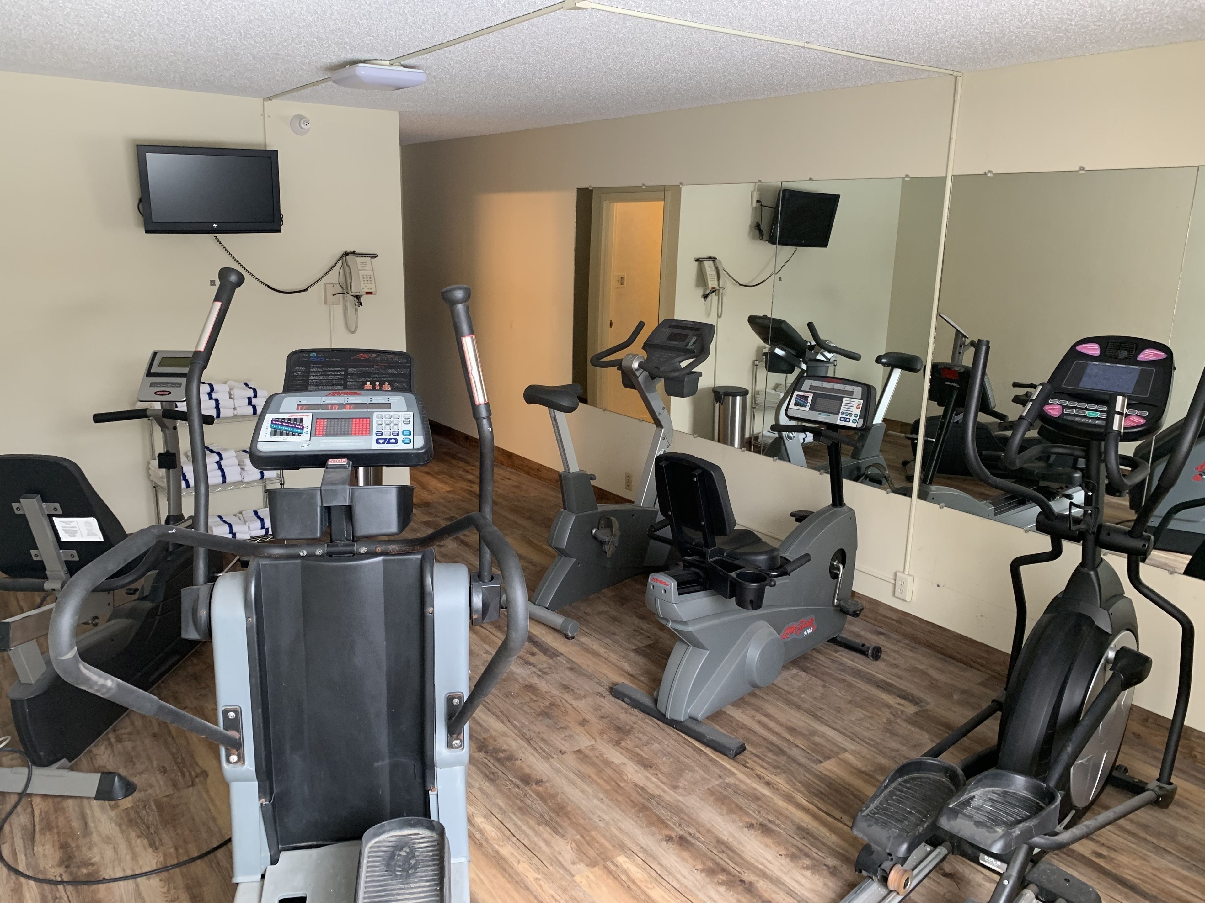 A gym with multiple exercise machines, a television mounted on the wall, and a large mirror reflecting the equipment.