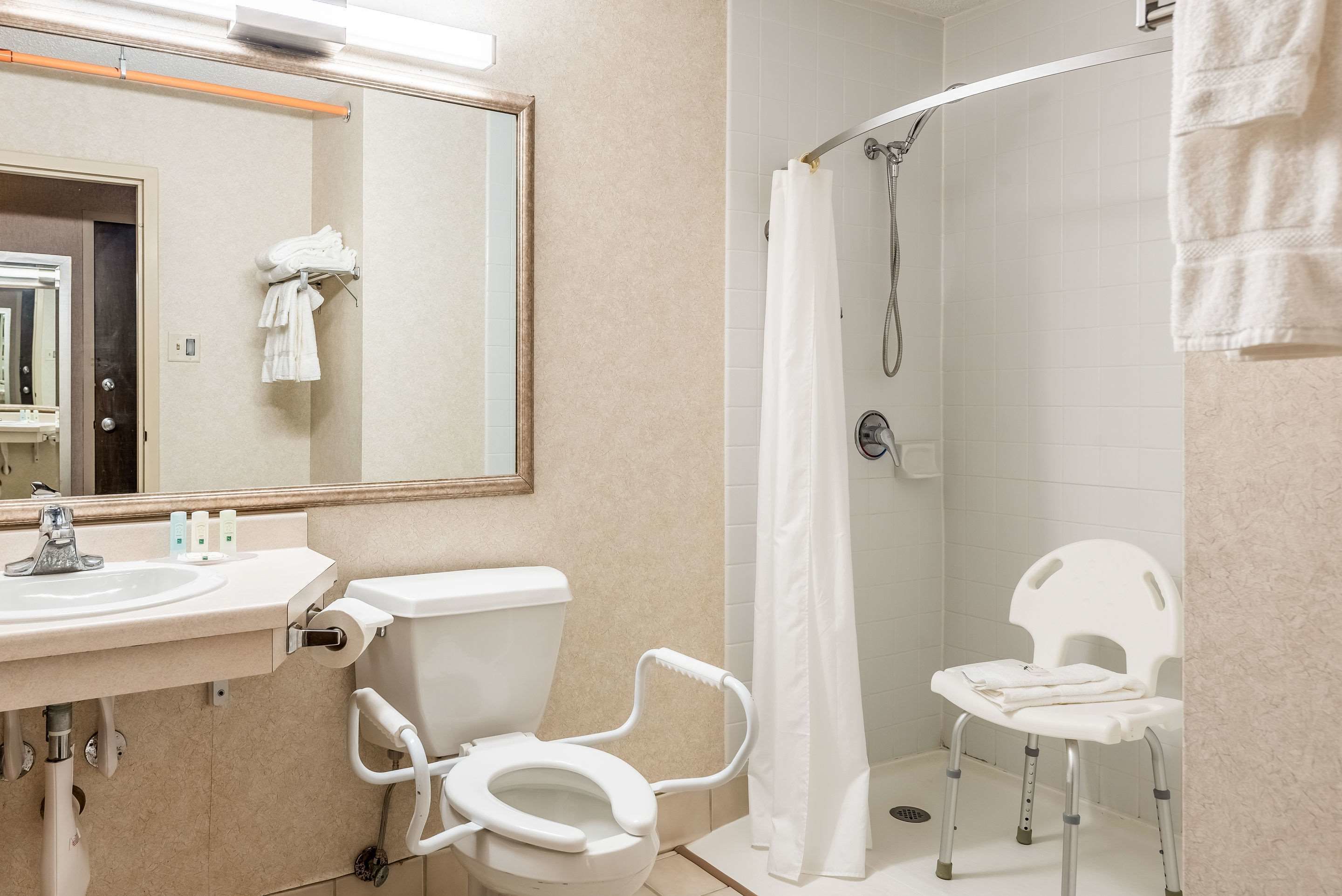 A bathroom with a toilet, sink, shower, and a white chair.