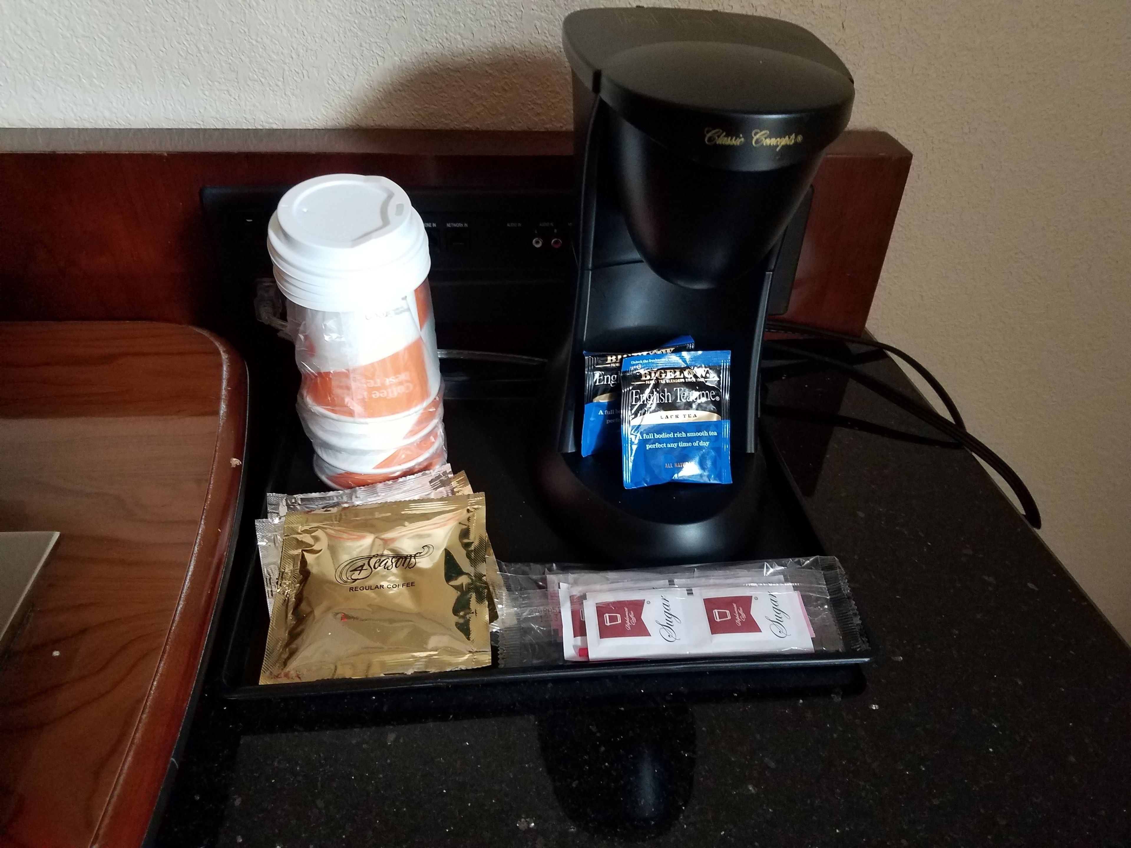 A coffee maker, a stack of disposable coffee cups, a bag of coffee pods, and a tray with sugar packets on a countertop.