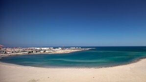 Eine Küstenlandschaft mit Sandstrand, ruhiger See und Industriegebäuden im Hintergrund.
