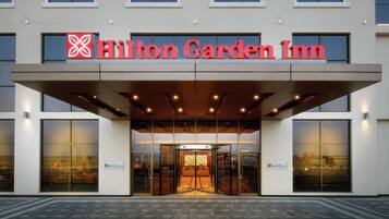 Exterior de um Hilton Garden Inn, com uma placa bem visível, grandes portas de vidro e um design de entrada moderno.