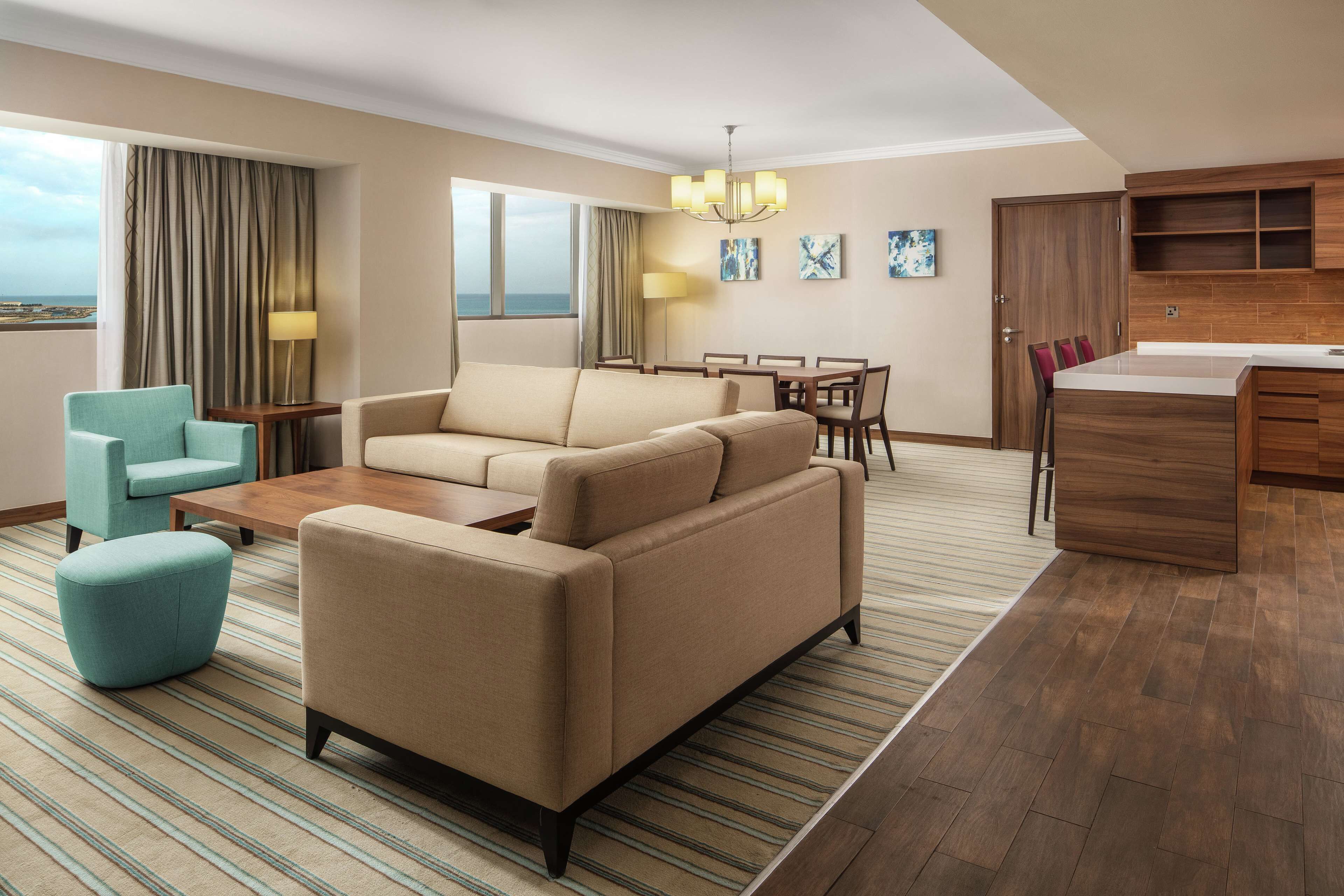 A modern hotel room with a sofa, ottoman, dining table, and chairs. There is a kitchen area with wooden cabinets and a view of the ocean through large windows.
