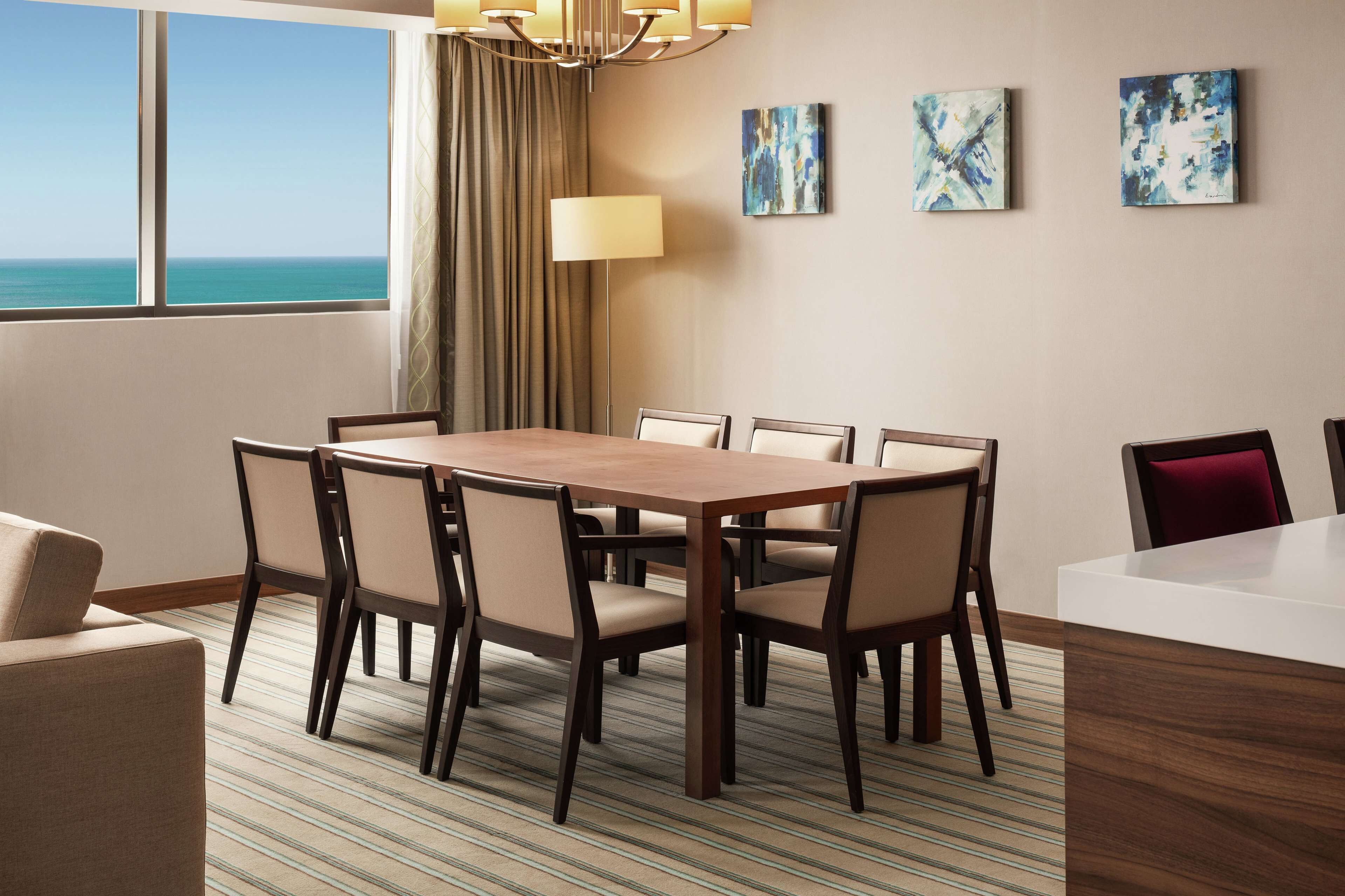 A hotel dining area with a large table, six chairs, a sofa, a lamp, and abstract art on the wall.