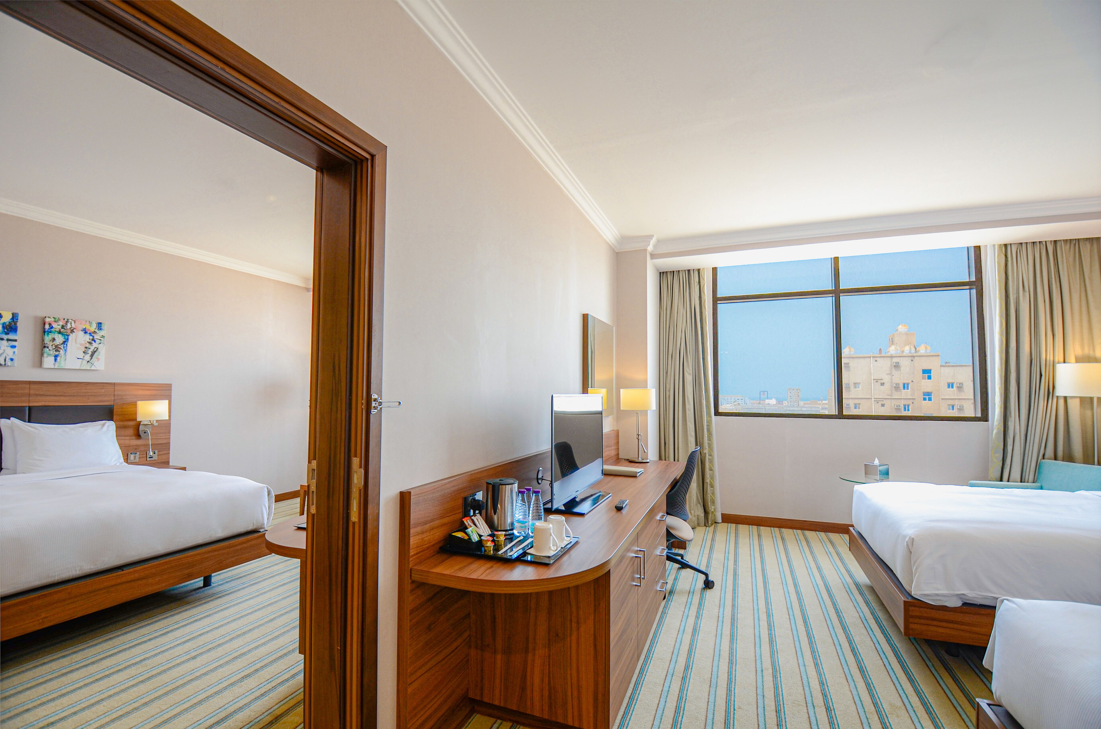 Ein Hotelzimmer mit zwei Betten, einem Schreibtisch mit Computer, einem Stuhl und Blick auf ein Gebäude durch das Fenster.