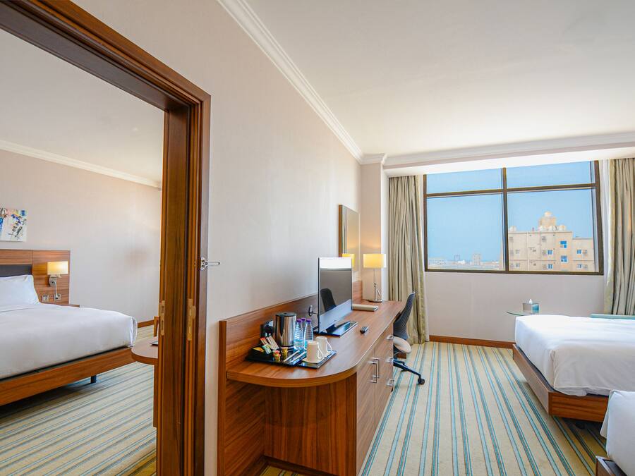 Ein Hotelzimmer mit zwei Betten, einem Schreibtisch mit Computer, einem Stuhl und Blick auf ein Gebäude durch das Fenster.
