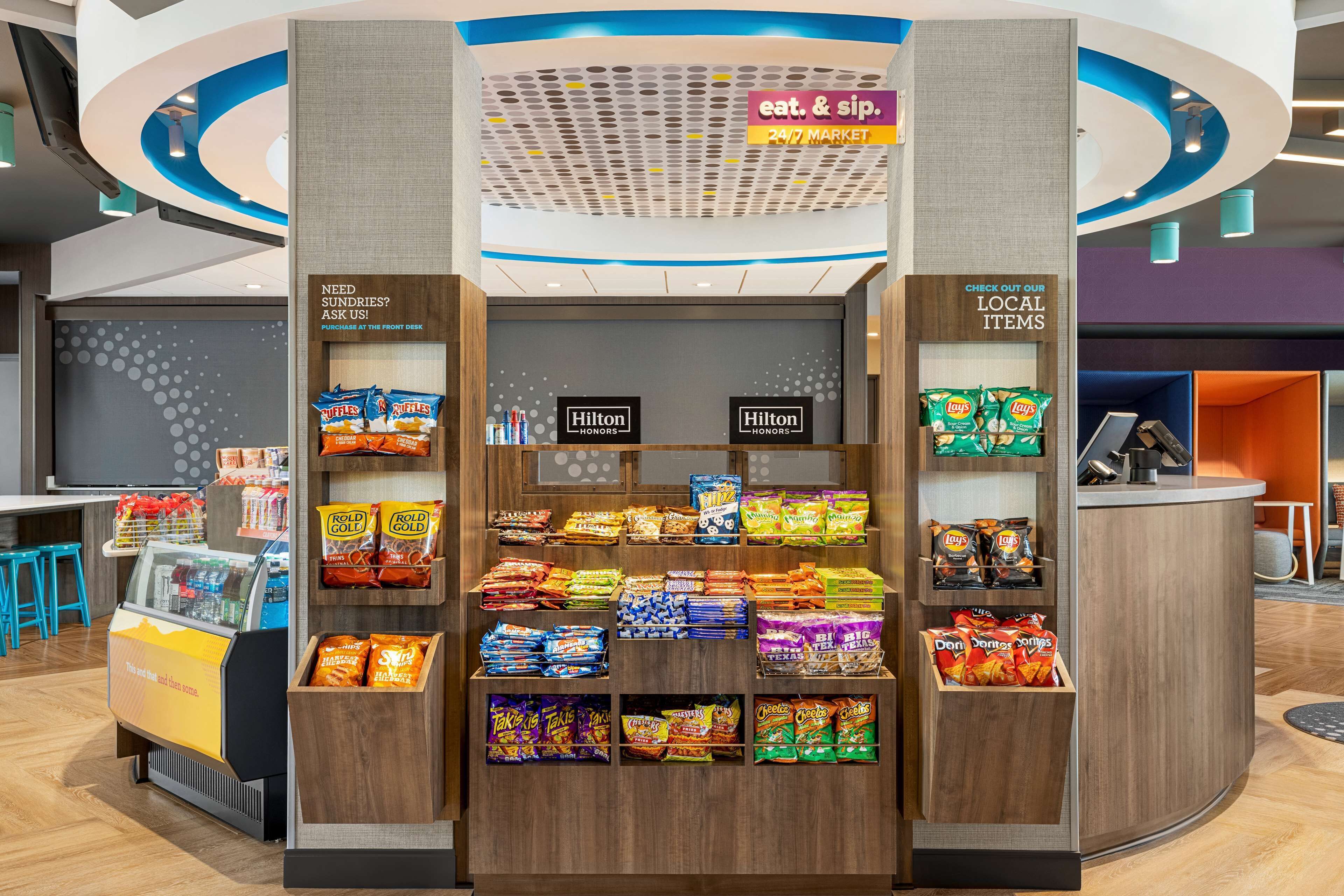 Un quiosc de snacks amb diverses marques com Ritz, Lay's i Doritos. Hi ha cartells que diuen 'menja i beu' i 'supermercat 24/7'.