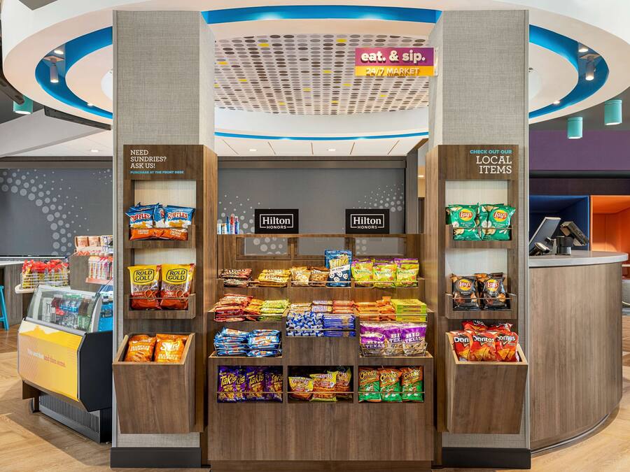 Un quiosc de snacks amb diverses marques com Ritz, Lay's i Doritos. Hi ha cartells que diuen 'menja i beu' i 'supermercat 24/7'.