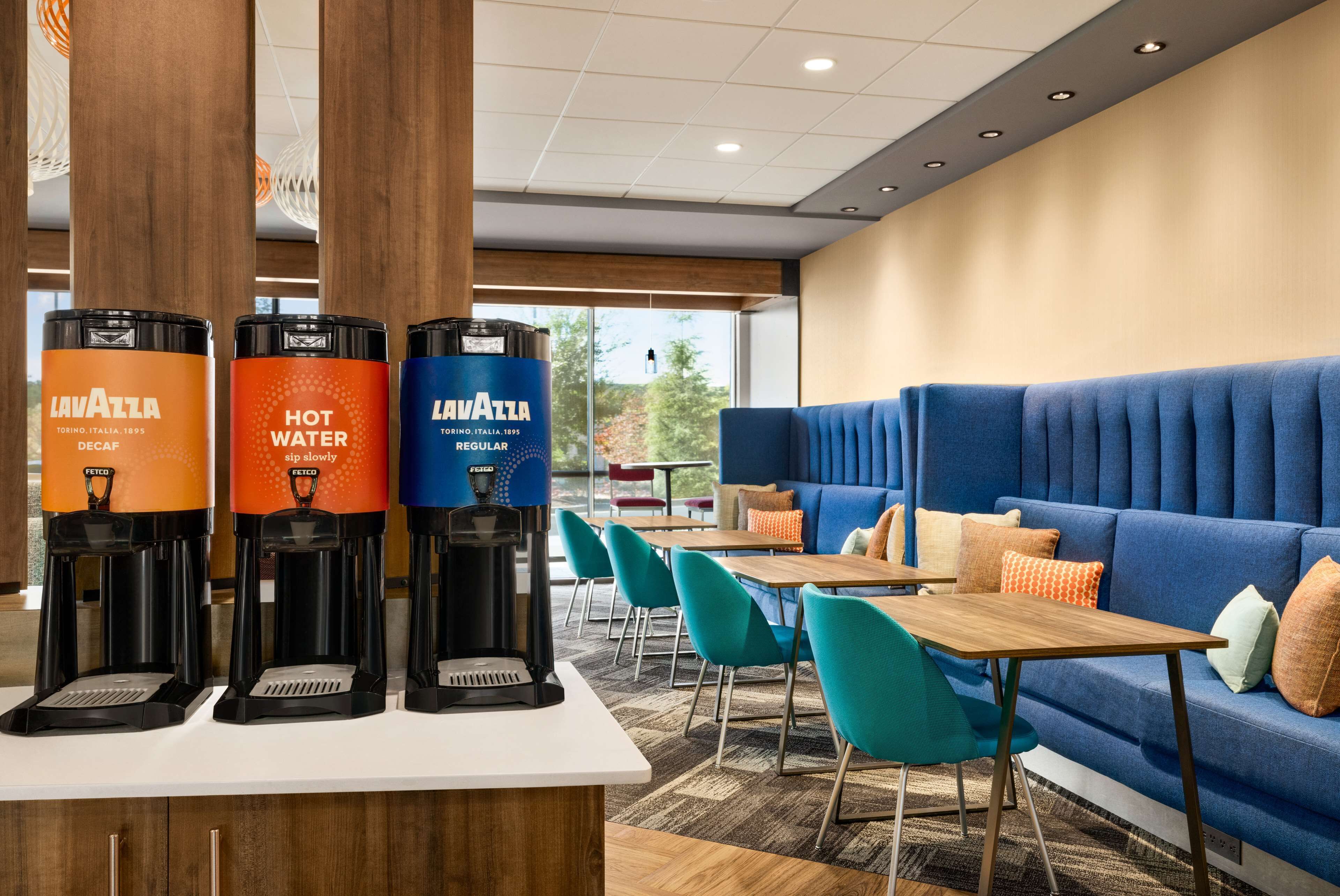 Un hall d’hôtel moderne doté d’un espace salon, d’un coin café avec trois distributeurs et offrant une vue sur l’extérieur.