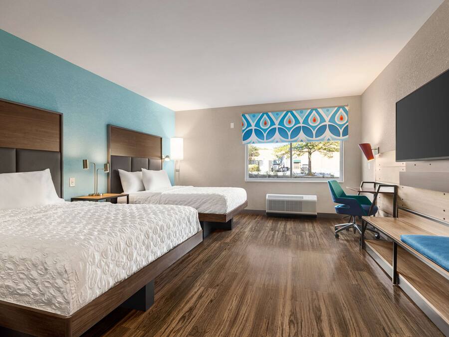 A modern hotel room with two beds, a desk, a chair, and a large window.