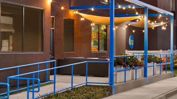 A covered outdoor seating area with string lights, a blue railing, and a building entrance with a logo.