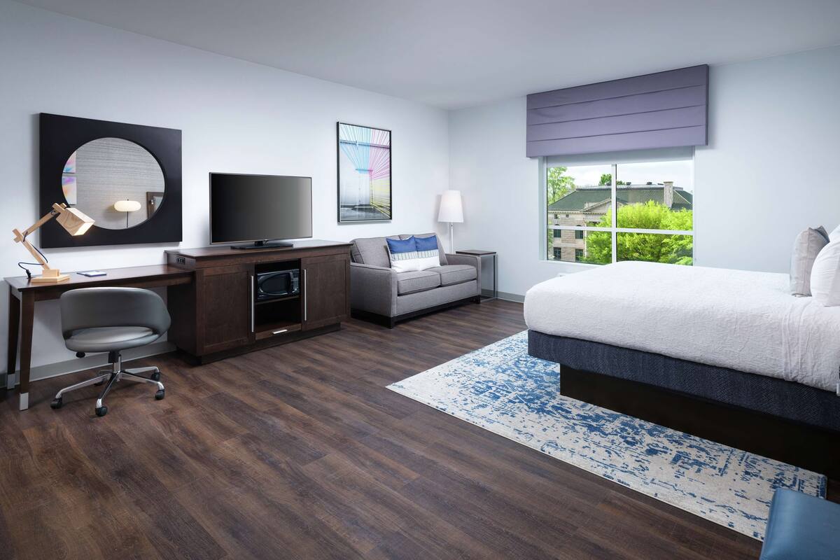 A modern hotel room with a large bed, a desk with a flat-screen TV, a comfortable sofa, and a view of the city through the window.
