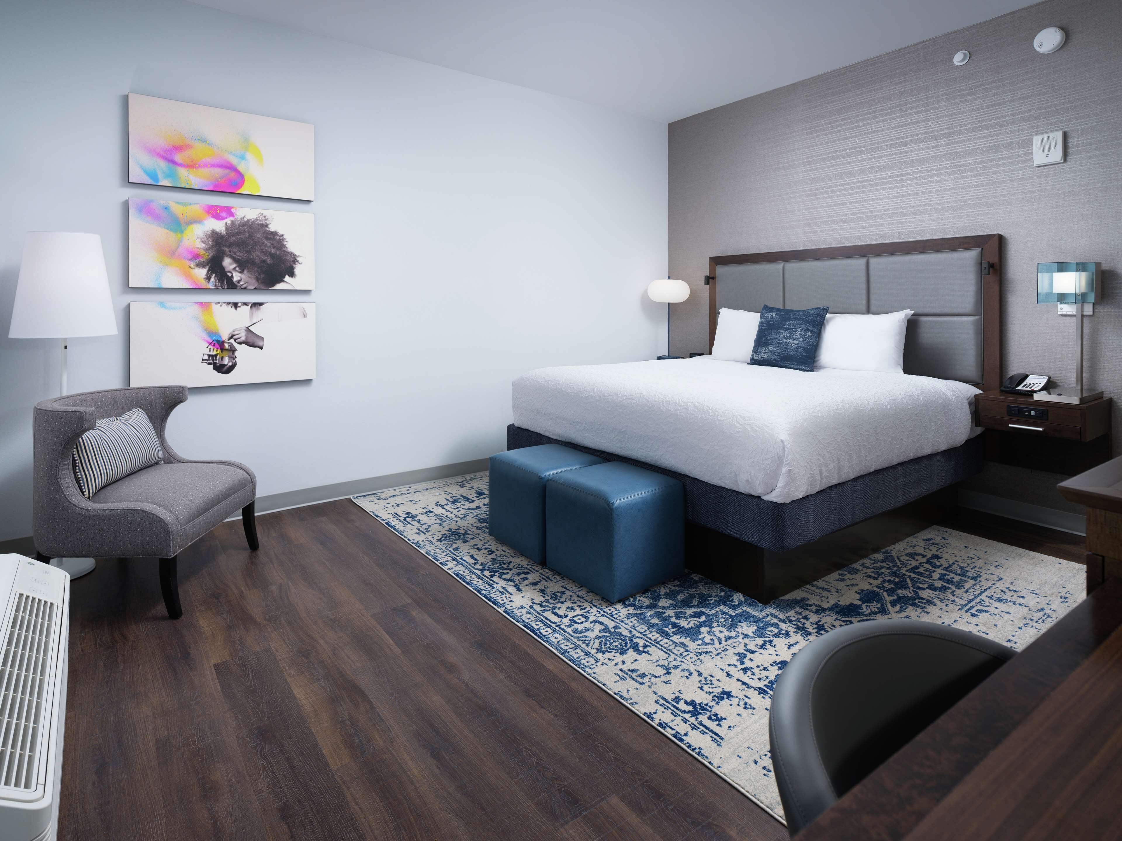 Habitación de hotel moderna con una cama grande, un sillón gris, un otoman azul, un escritorio con lámpara y un teléfono en la pared.