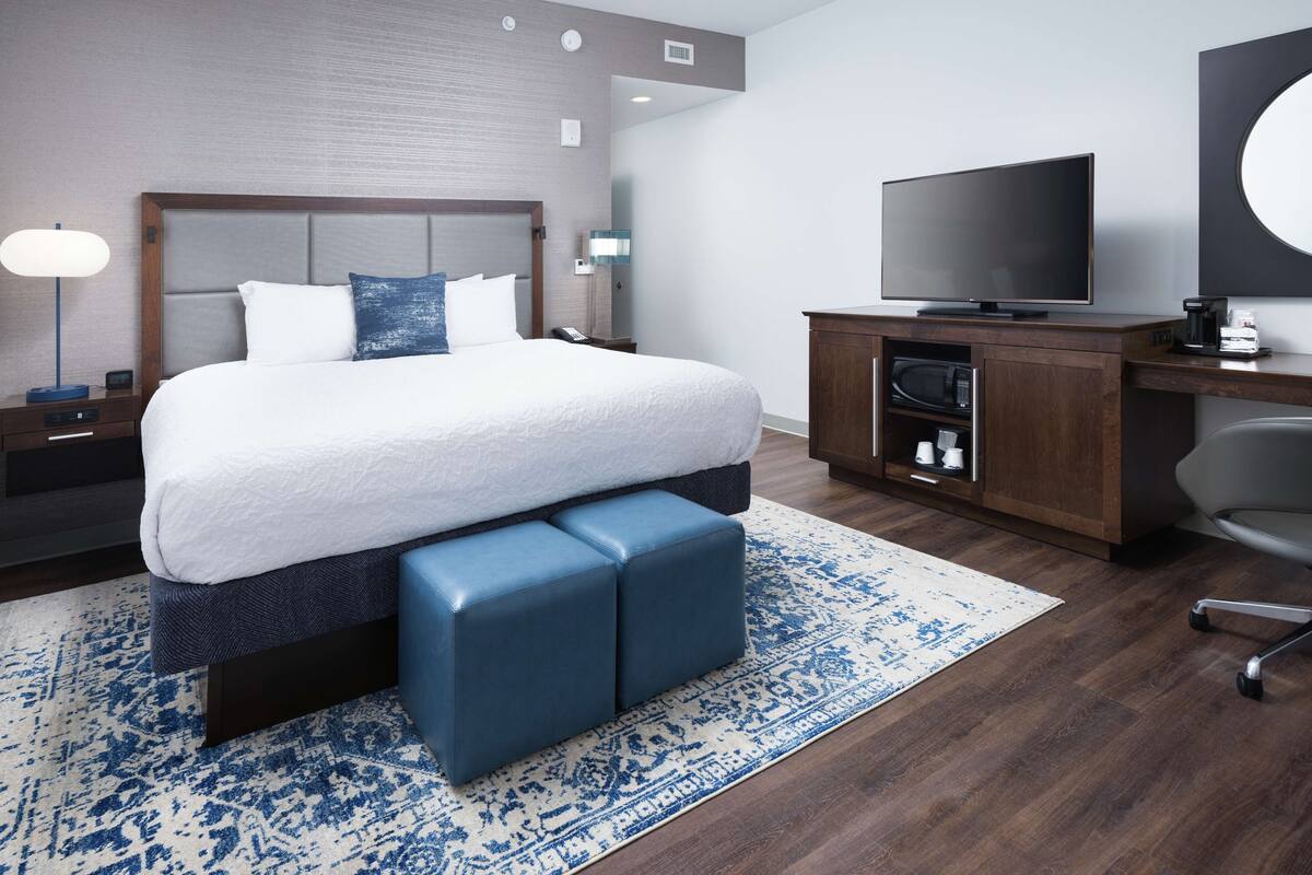 A modern hotel room with a large bed, a flat-screen TV, a wooden desk, and a small kitchenette.