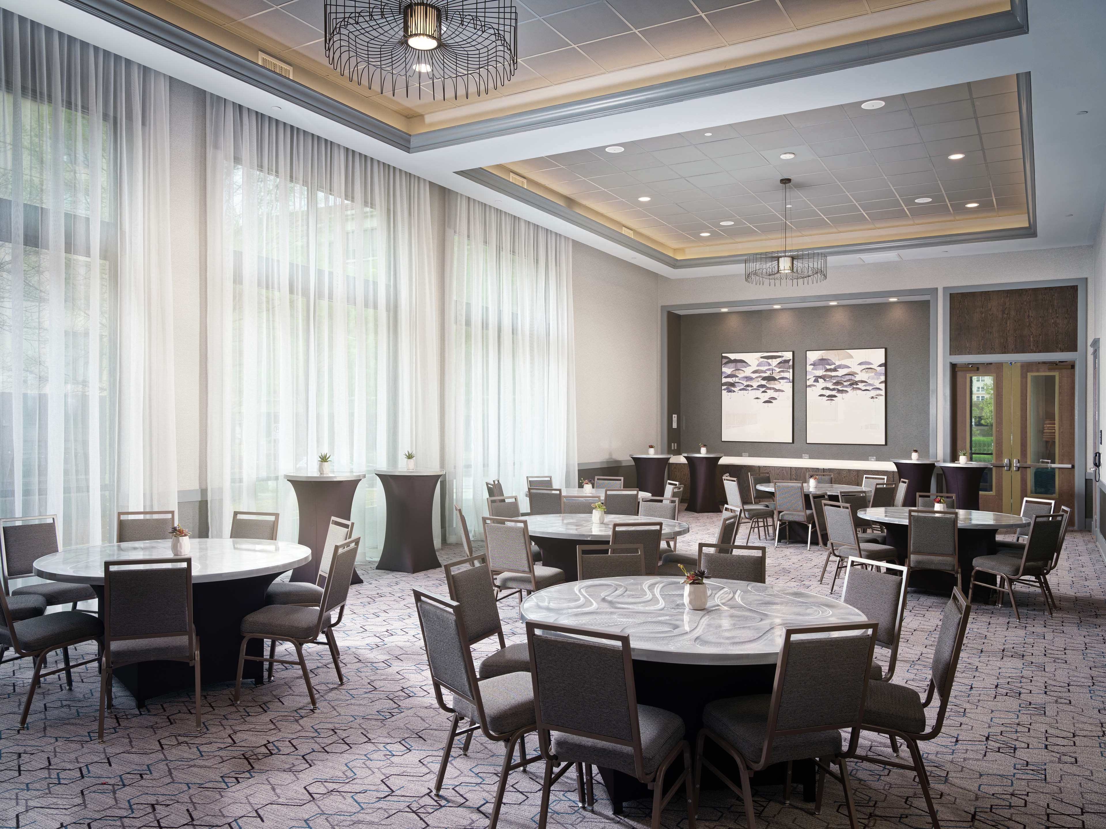 A spacious conference room with round tables and chairs, large windows with sheer curtains, and a modern ceiling design.