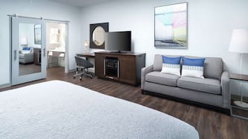 A modern hotel room with a large bed, a gray sofa with blue pillows, a desk with a computer and lamp, a TV, and a sliding door leading to a bathroom.