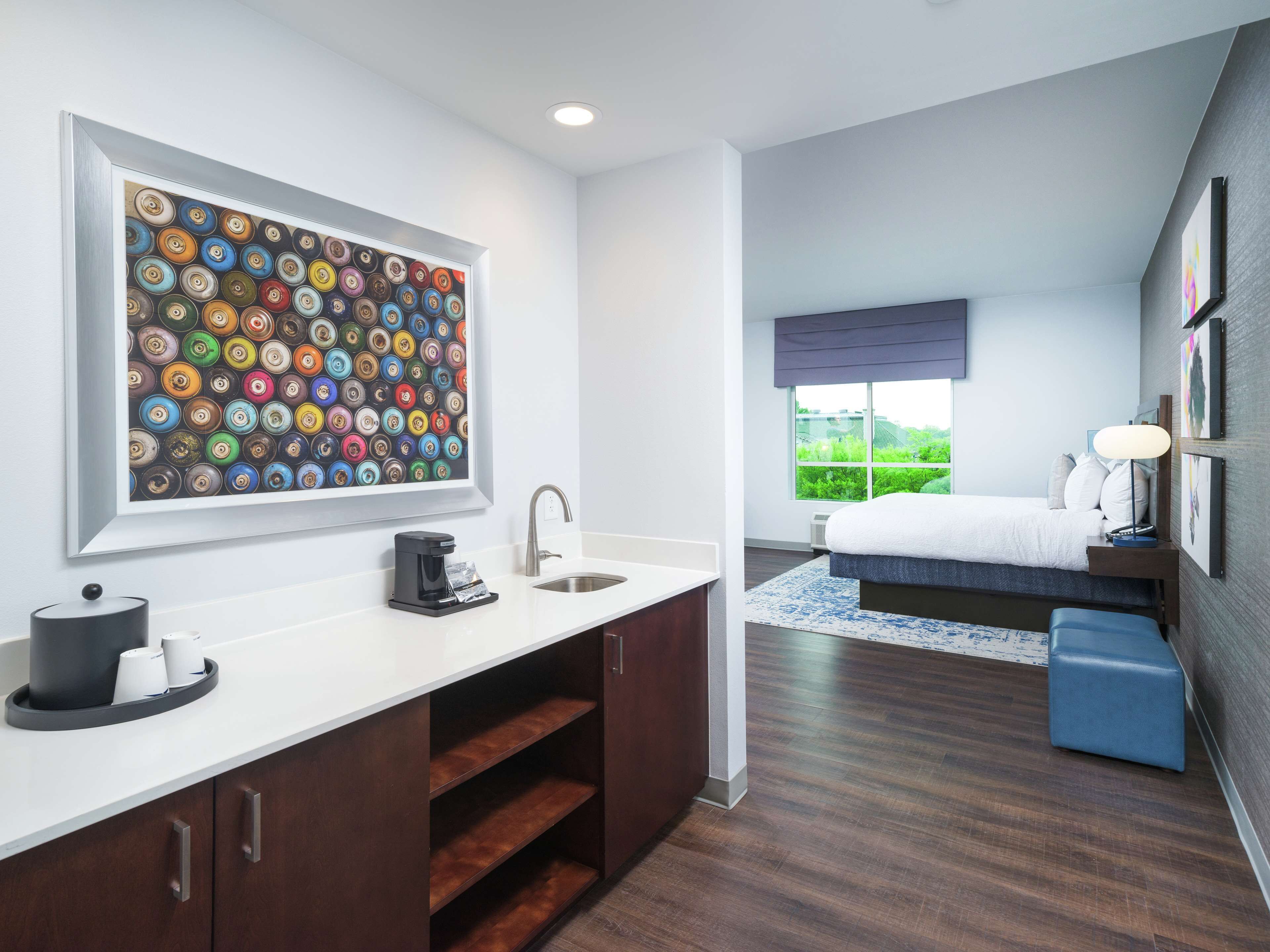 A modern hotel room with a large wall art of colorful circular patterns, a built-in kitchen with a coffee maker, a sink, and a bedside table with a lamp.