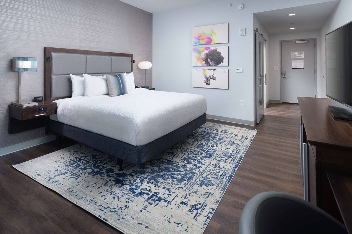 A modern hotel room with a large bed, a desk, and a chair. There are two bedside tables with lamps and a wall-mounted TV. The room has a wooden floor and a patterned rug.