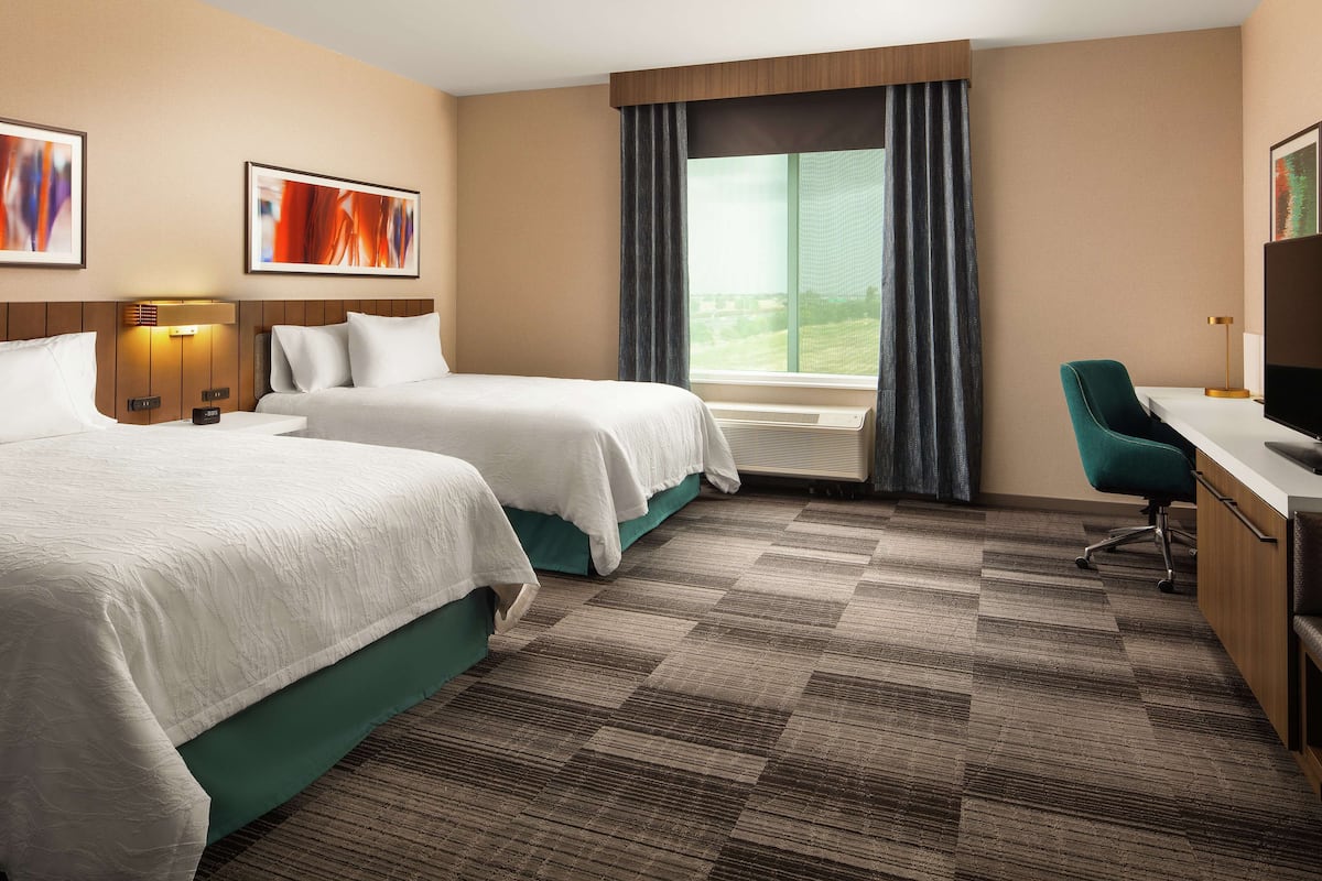 A hotel room with two beds, a desk with a chair, a TV, and a large window with curtains.