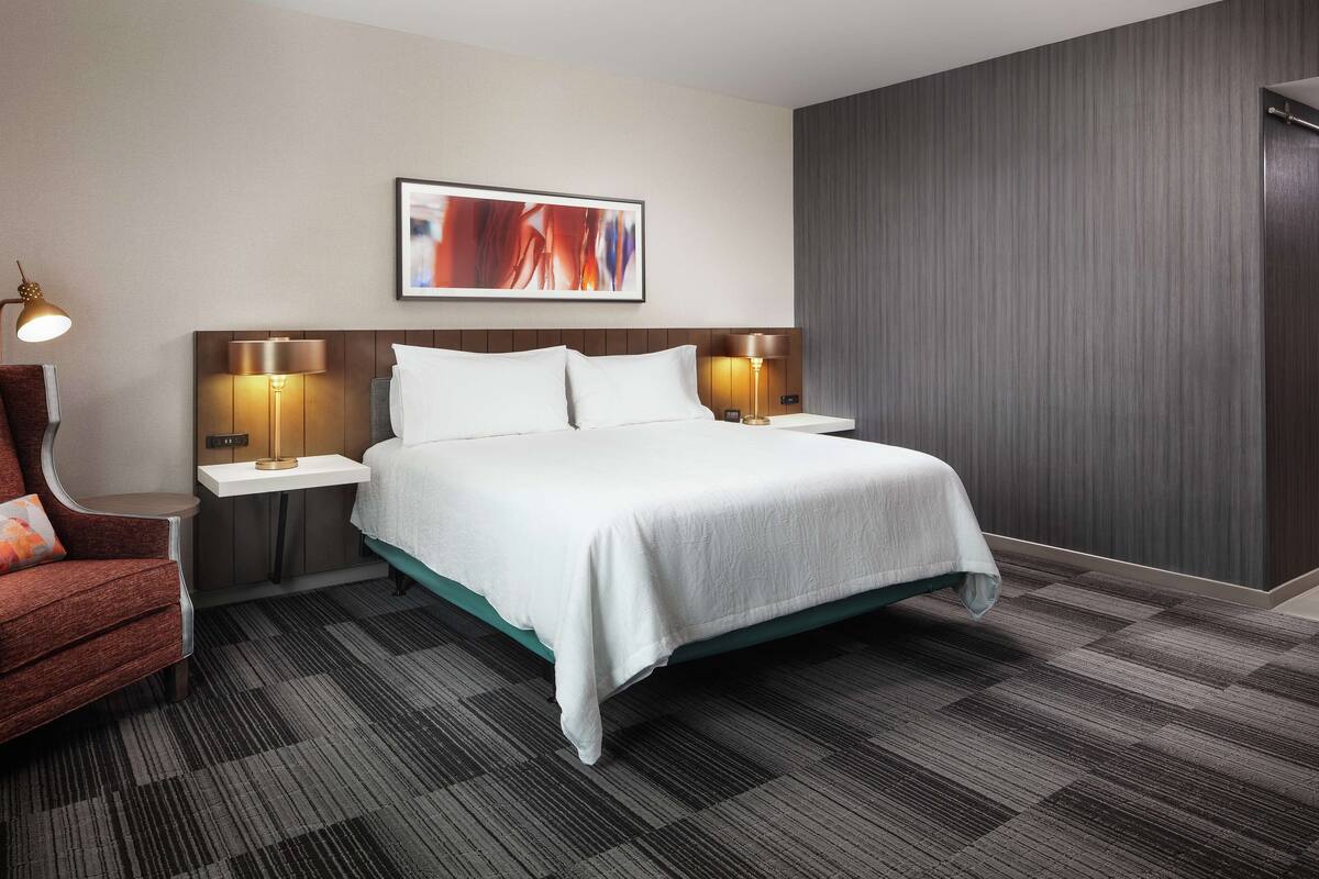 A hotel room with a bed, two bedside tables with lamps, a chair, and a large abstract painting on the wall.