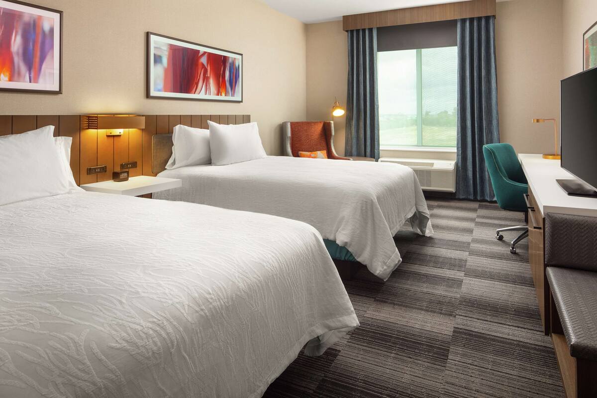 A hotel room with two beds, a desk, a chair, a TV, and a window with curtains.
