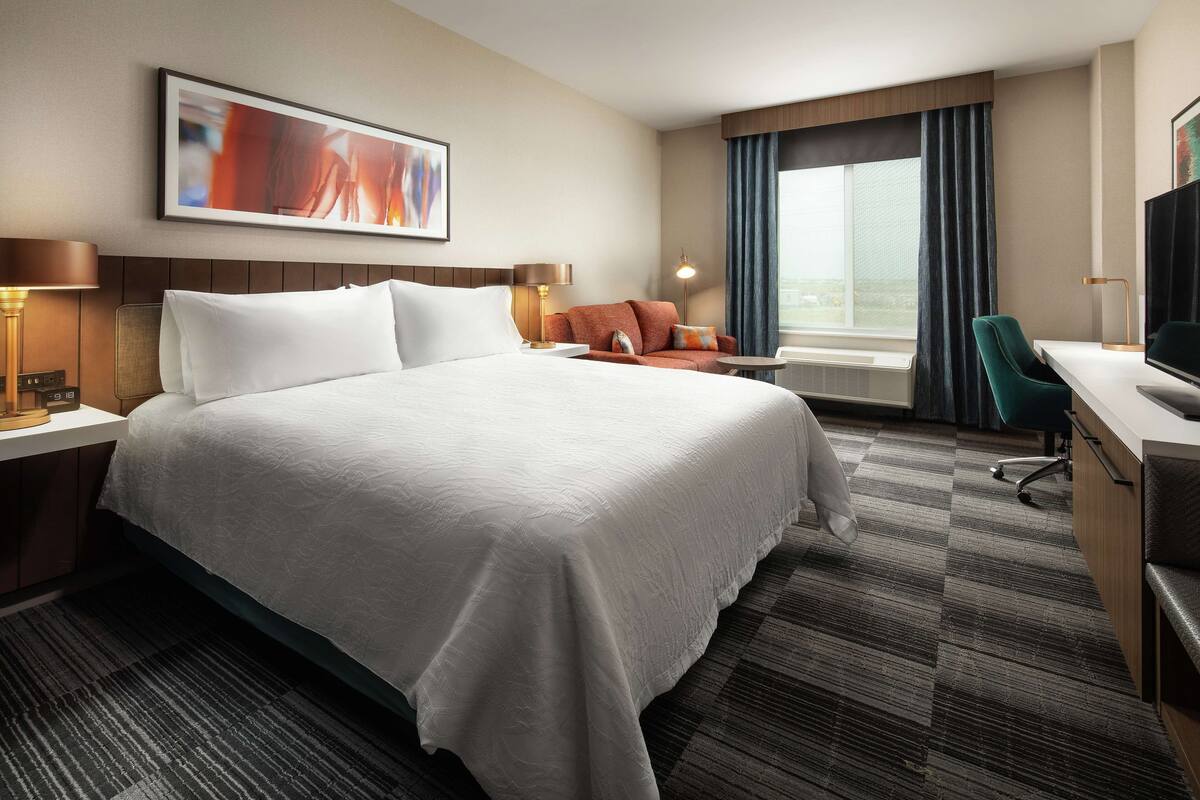A hotel room with a large bed, a desk, a chair, a TV, and a window with curtains.