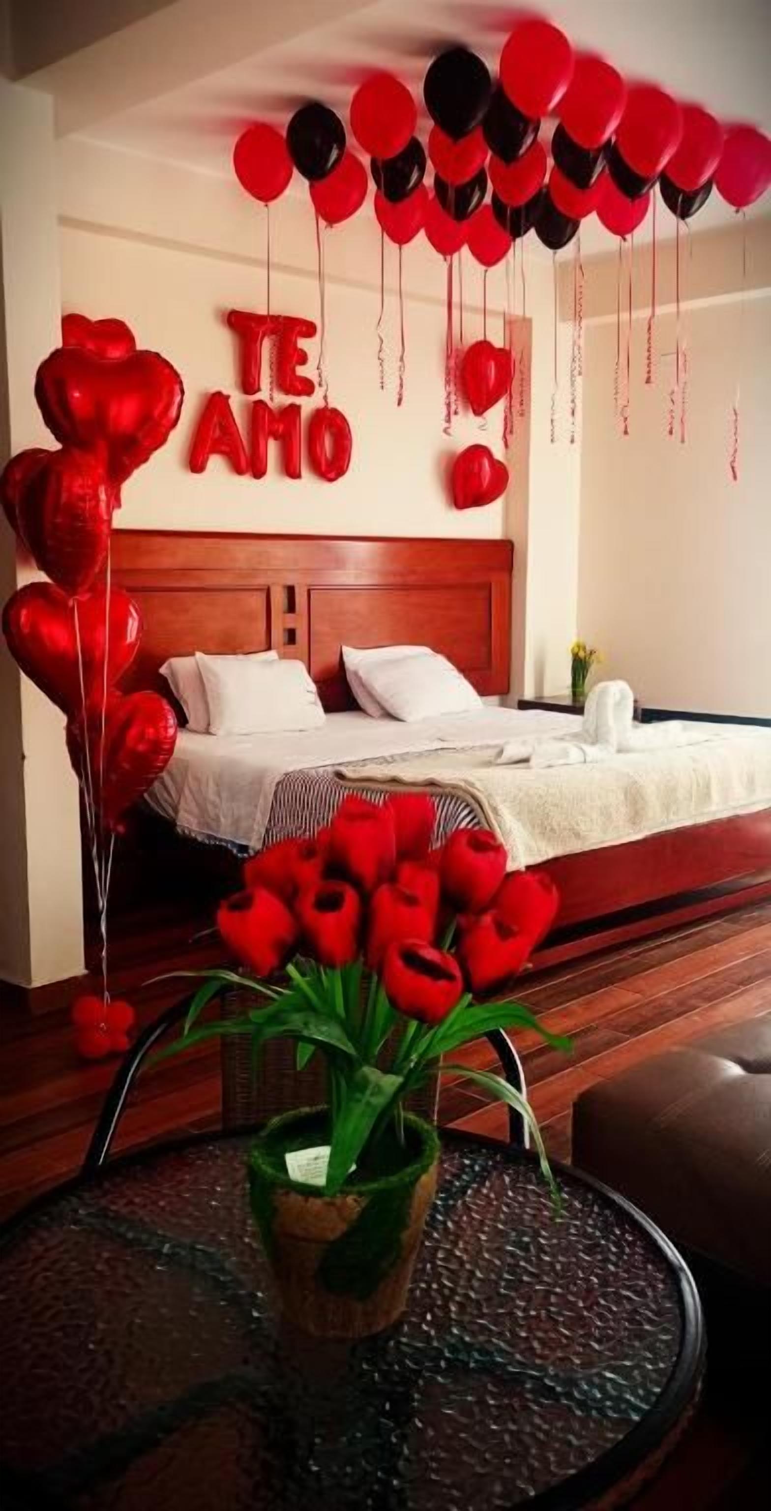 Une chambre avec un lit, une table de chevet ornée d’un vase de fleurs rouges, et un mur décoré de ballons rouges et noirs, sur lequel on peut lire le texte « TE AMO ».