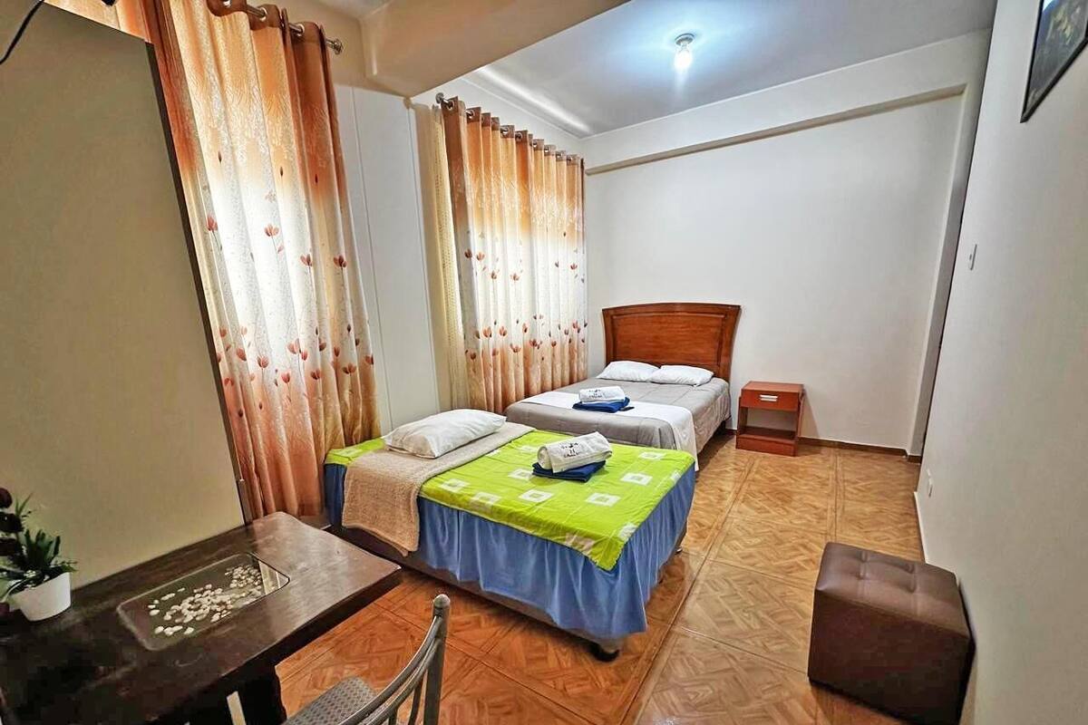 A small, well-lit room with two beds, a wooden floor, a desk with a chair, and a small table with a plant.