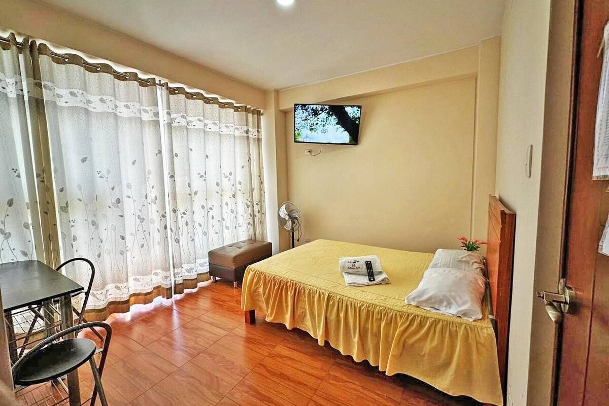 A hotel room with a bed, a TV mounted on the wall, a dining table with chairs, and a window with curtains.
