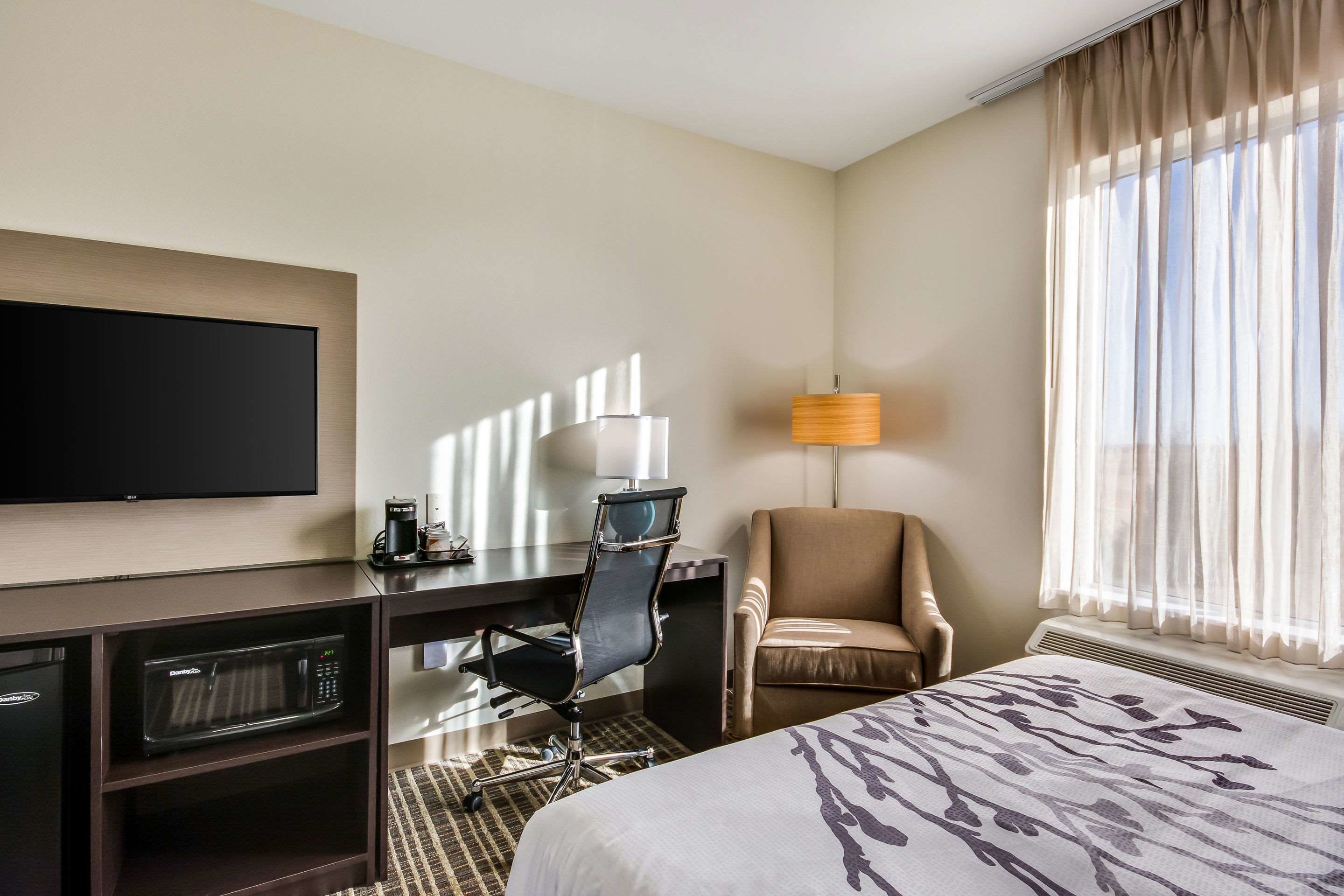 A hotel room with a bed, a desk with a computer, a chair, a TV, and a window with curtains.